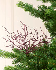 A close-up of a Christmas tree branch ornament with a Purple  twig-like branching pattern, nestled among the green needles of a pine tree against a light cream panelled wall.