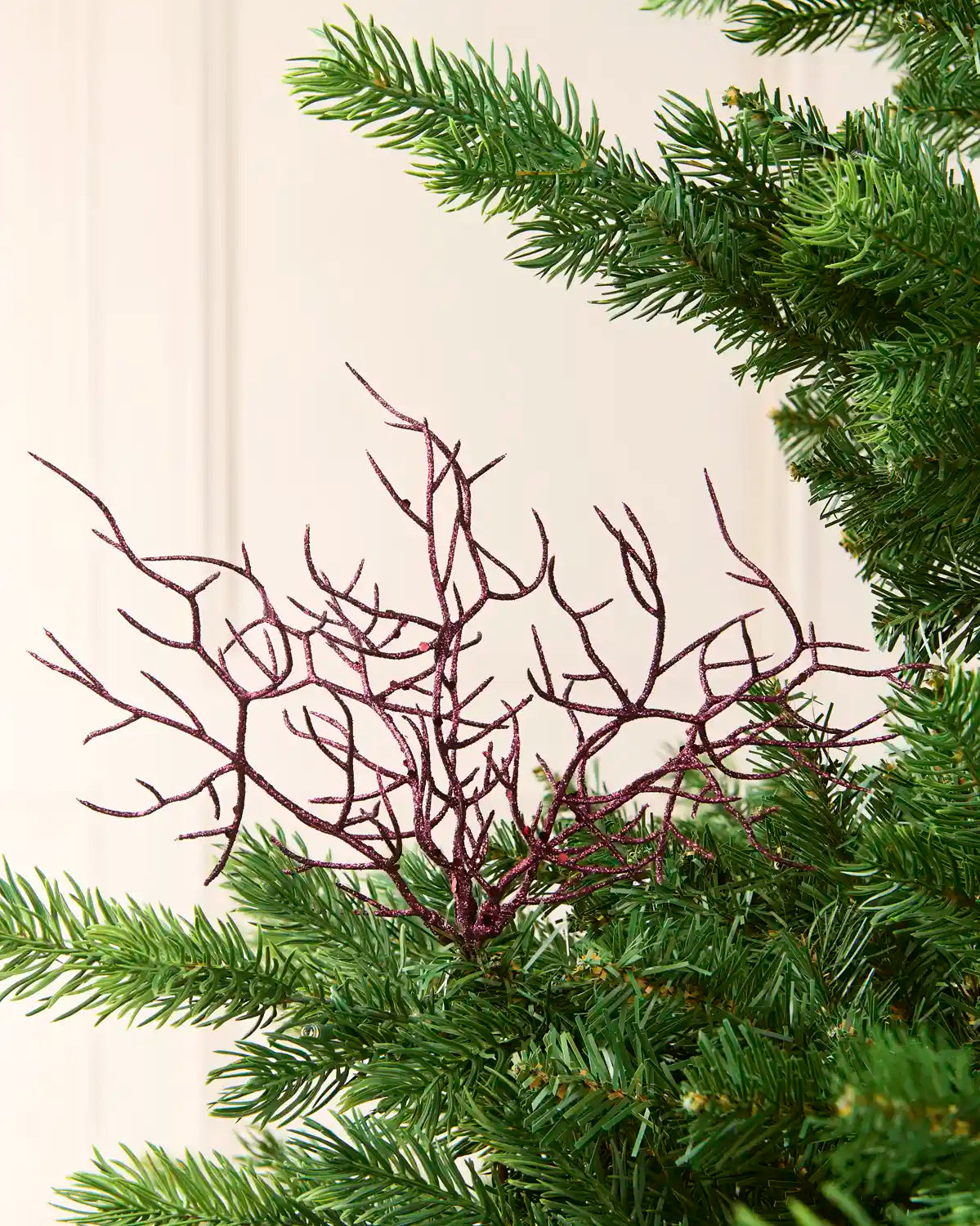 A close-up of a Christmas tree branch ornament with a Purple  twig-like branching pattern, nestled among the green needles of a pine tree against a light cream panelled wall.