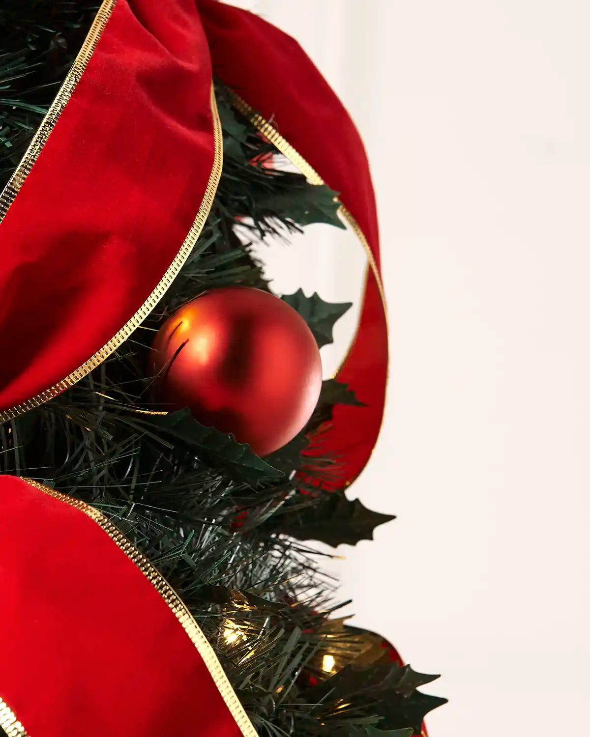 A close-up of a Christmas tree decorated with a large red velvet ribbon with gold trim, a shiny red bauble ornament, and green artificial holly leaves against a plain white background.