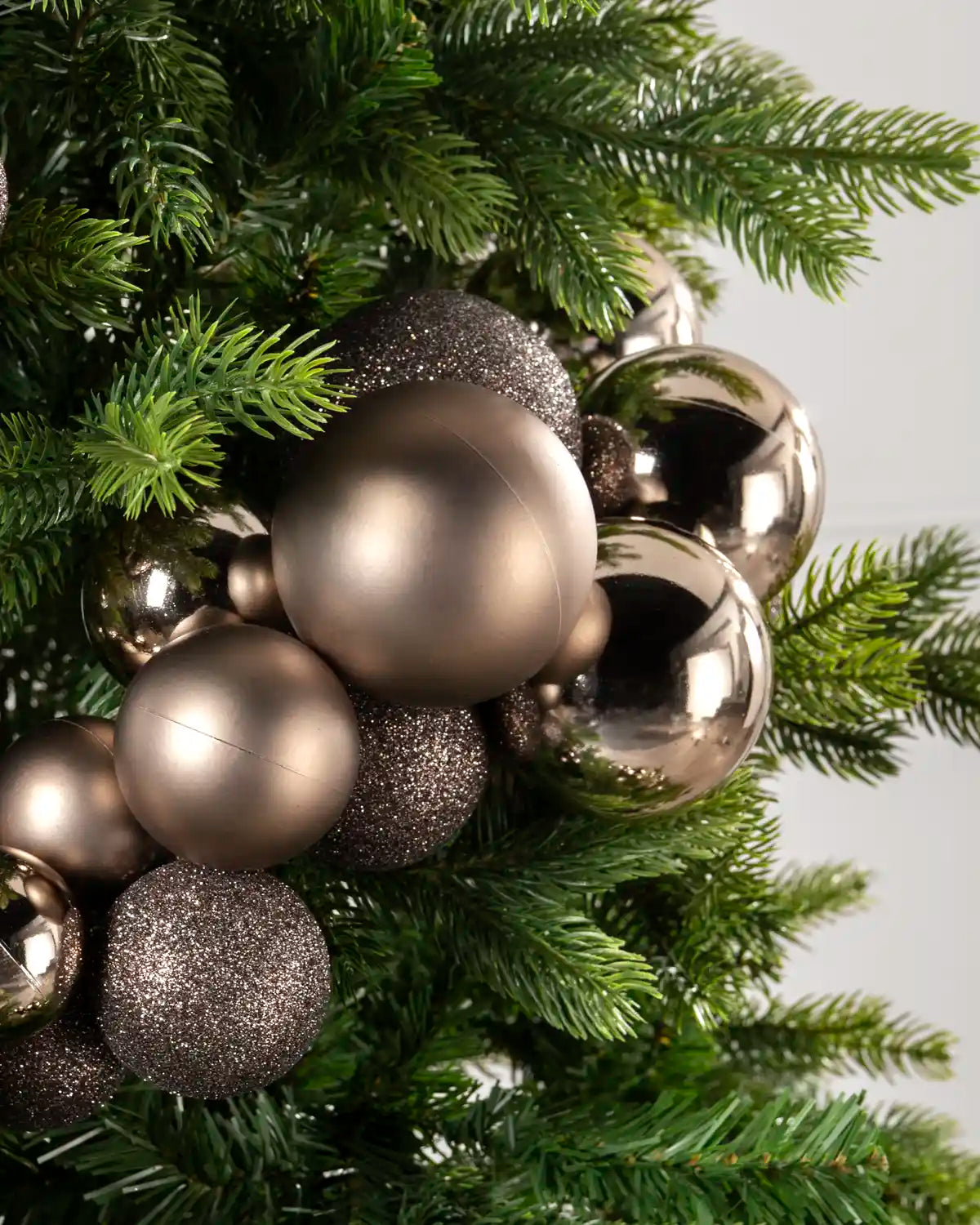 A close-up of a Christmas tree decorated with metallic and glittery Mocha ornaments in matte, brushed, and reflective finishes, nestled among green pine branches against a light gray background.