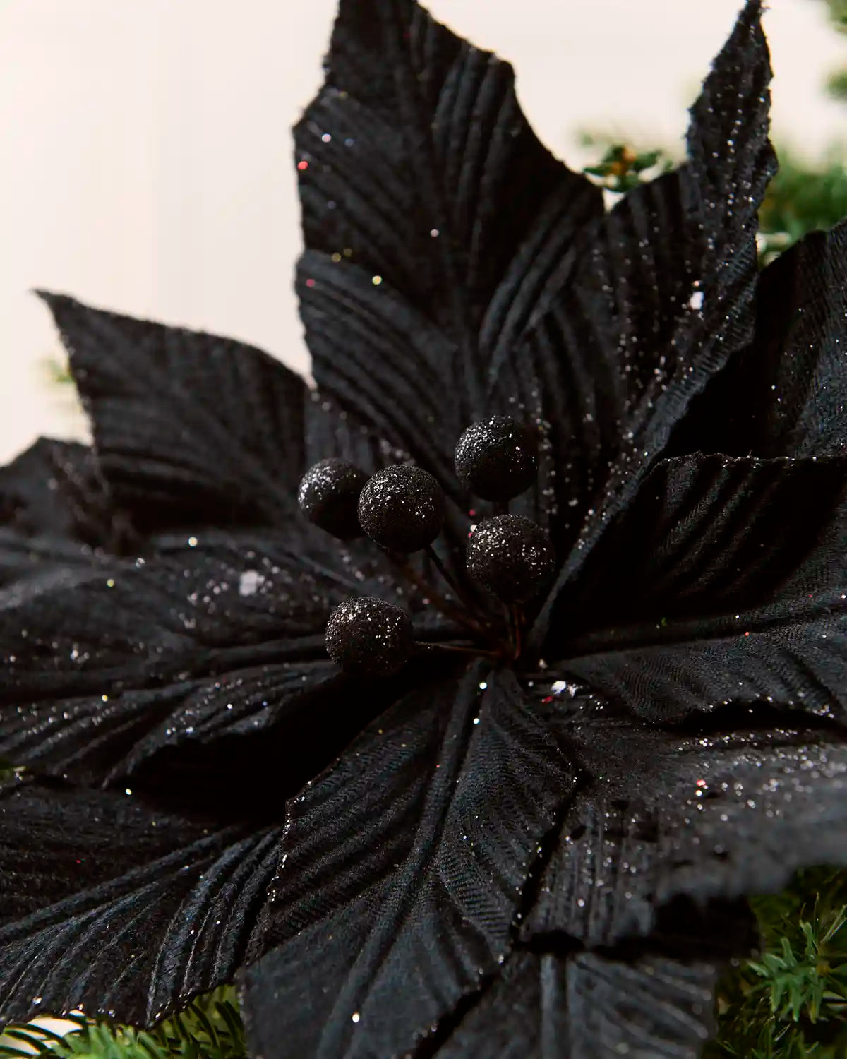 A close-up of a black artificial poinsettia decoration with textured, ribbed leaves and small glittery black berries, shown against a light background with green foliage at the base.