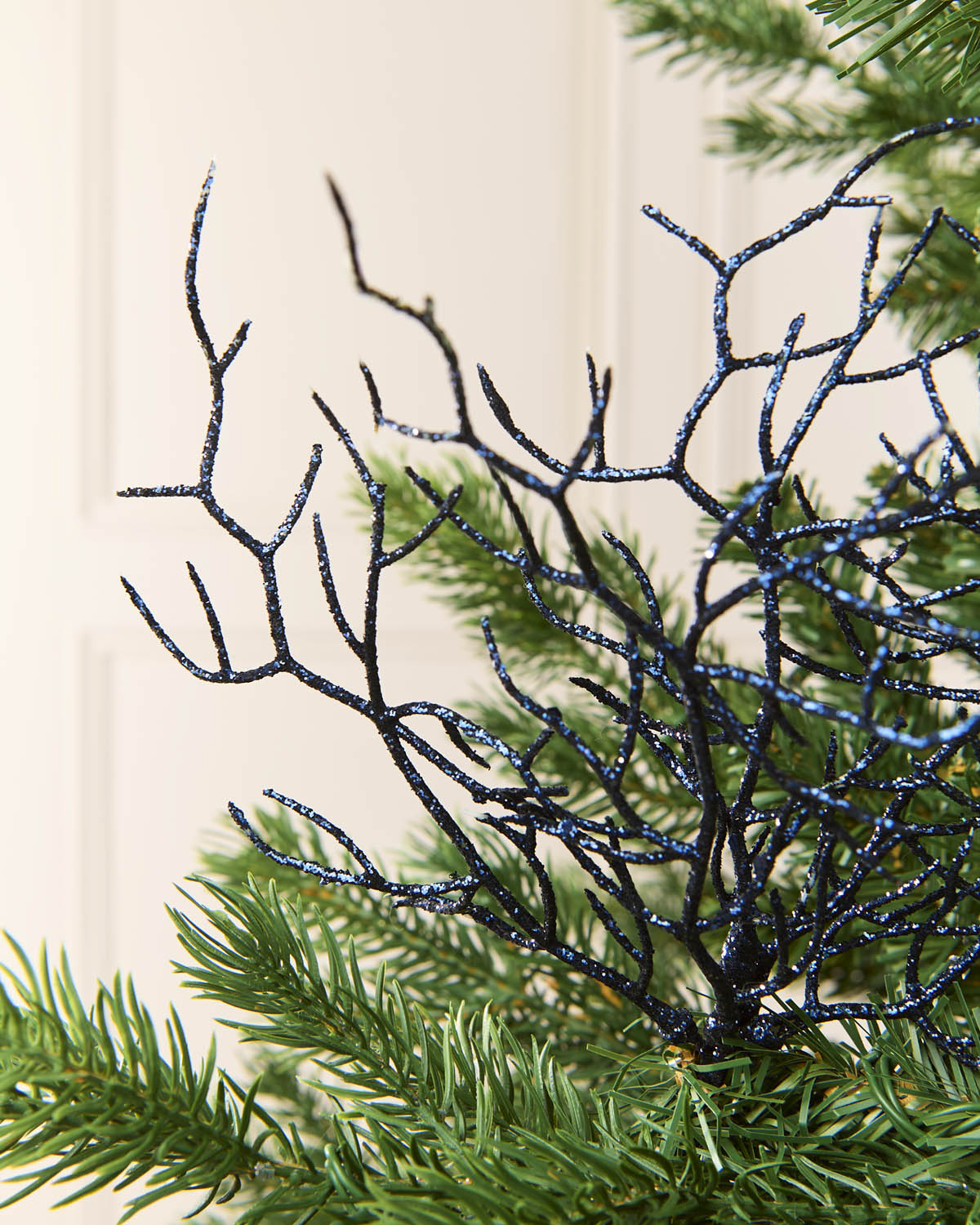 A close-up of a black glittery coral style decorative piece layered over green pine needles, with a soft cream paneled background visible behind it.