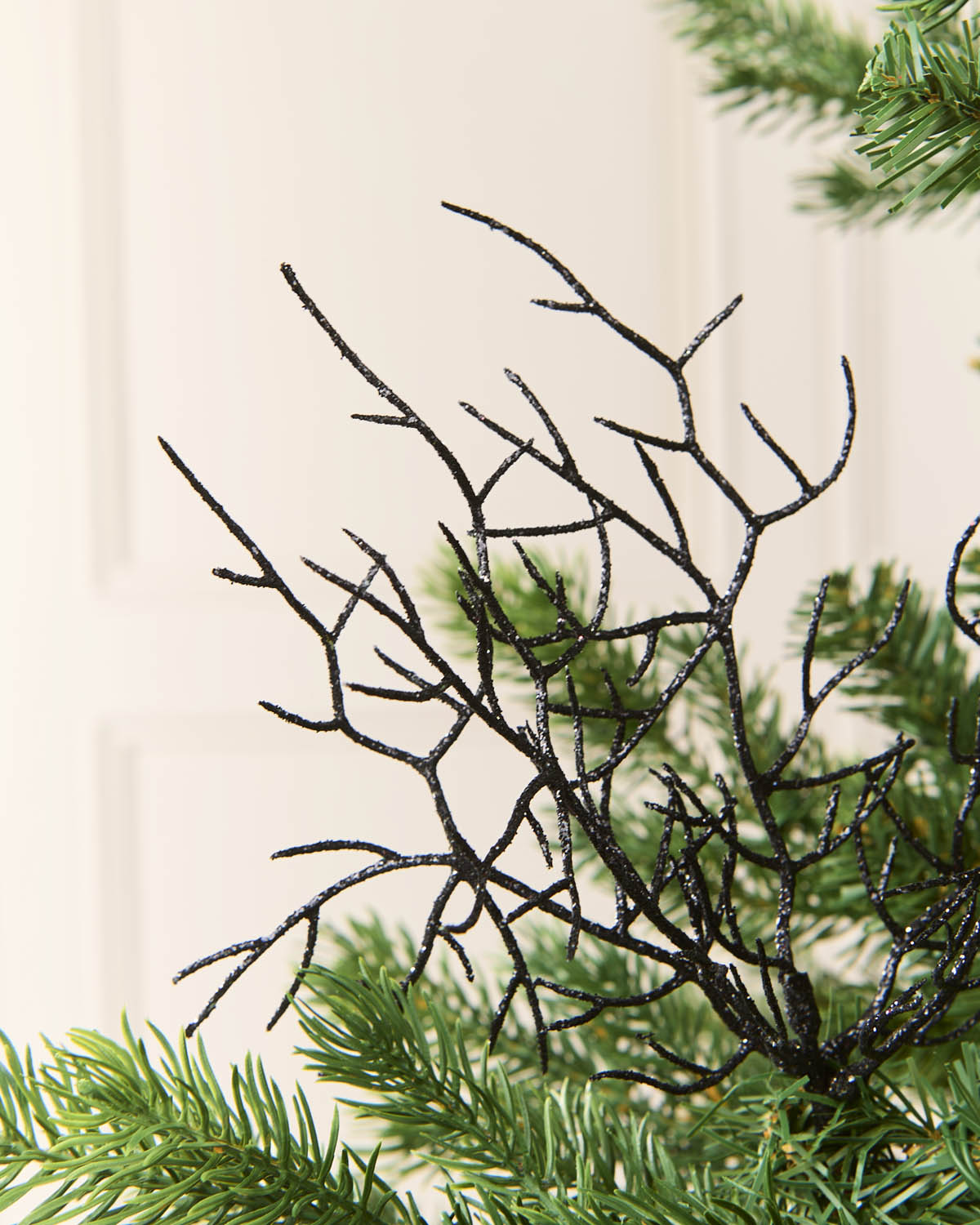 A close-up of a black glittery coral style  twig structure layered over green pine needles, against a soft cream-colored paneled background.