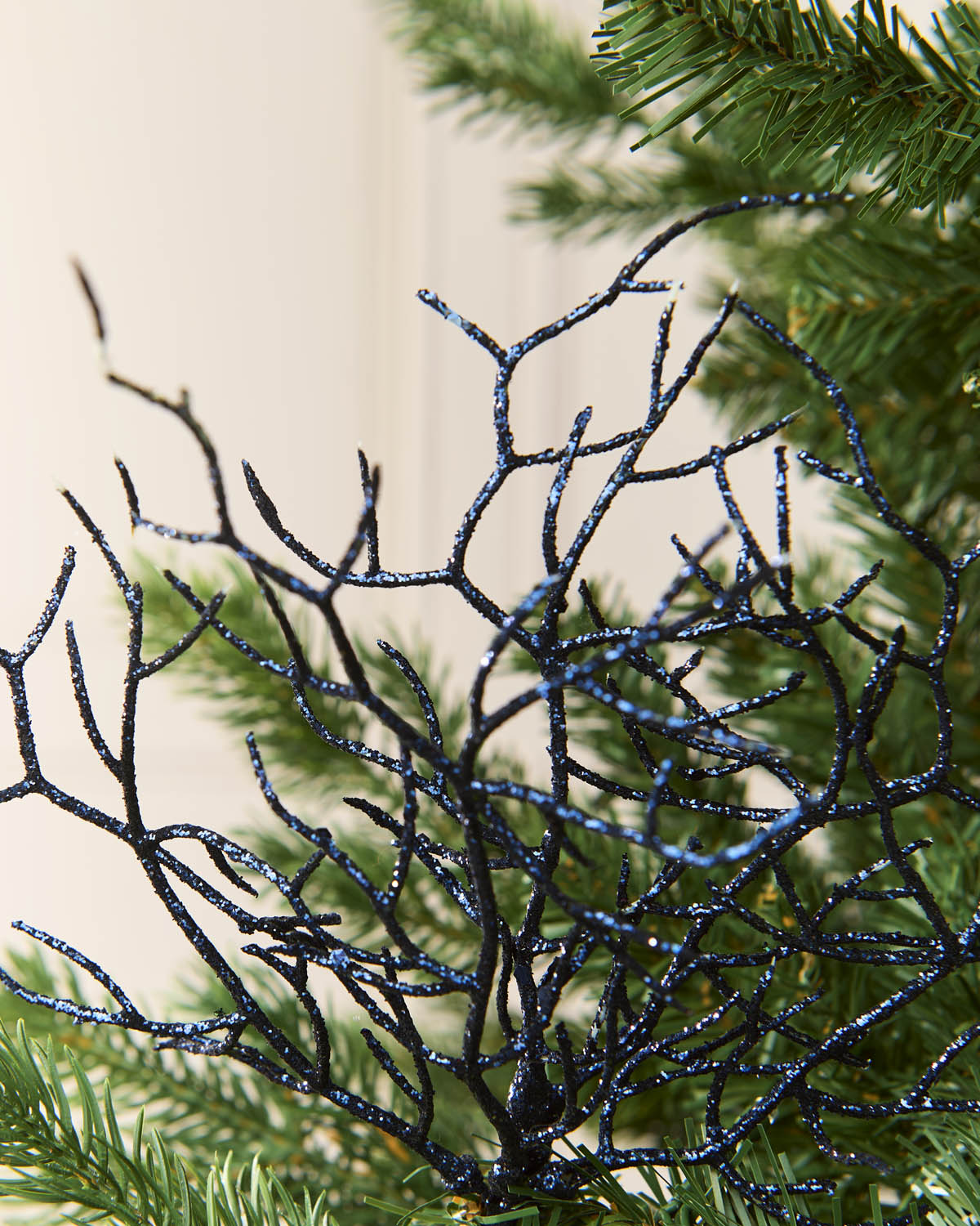 A close-up of a black glittery coral style twig structure with fine glitter strands, shown in front of green pine needles against a soft cream background.