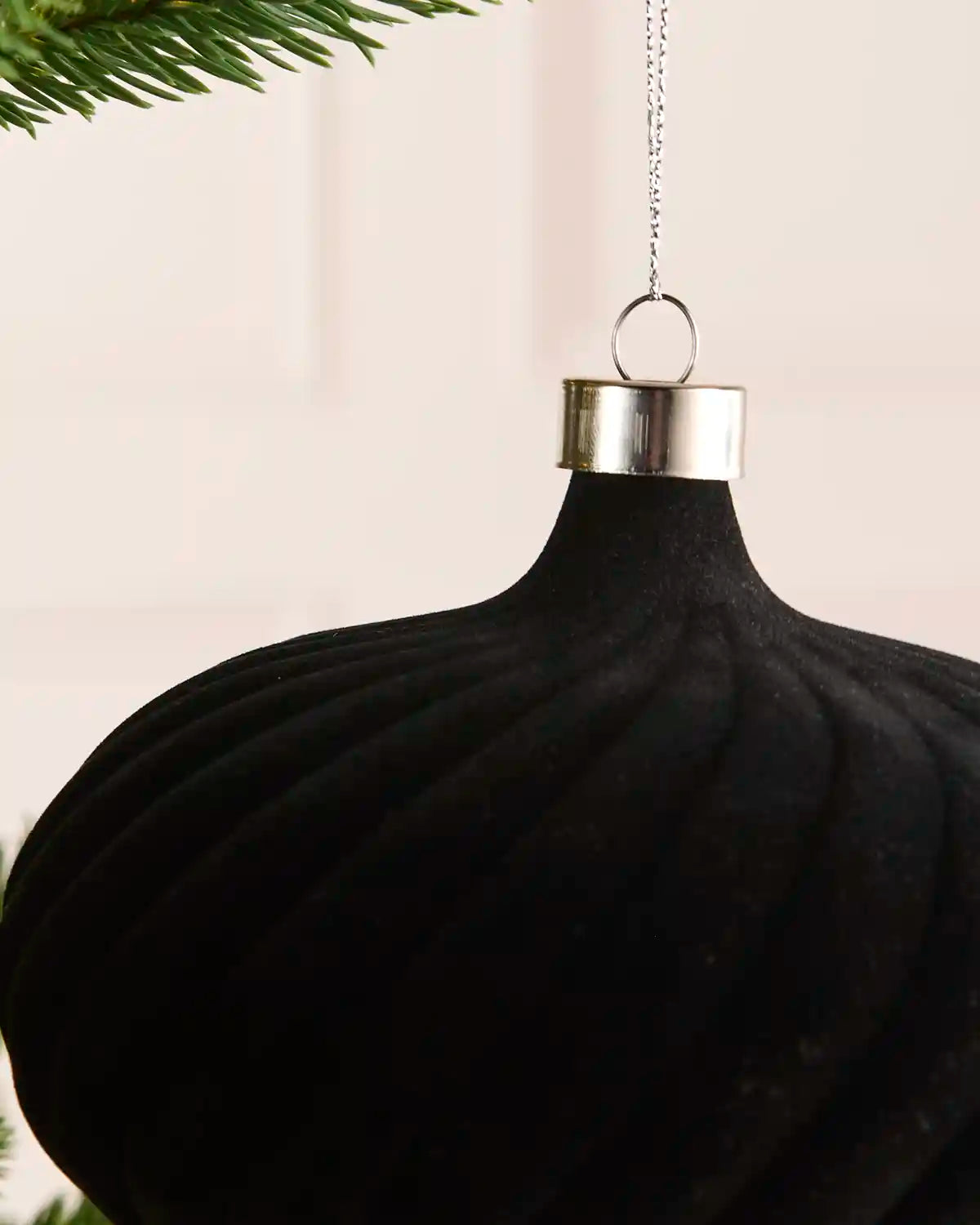 A close-up of a black ribbed Christmas ornament hanging from a silver cap with a silver chain, with green pine needles visible at the top left.