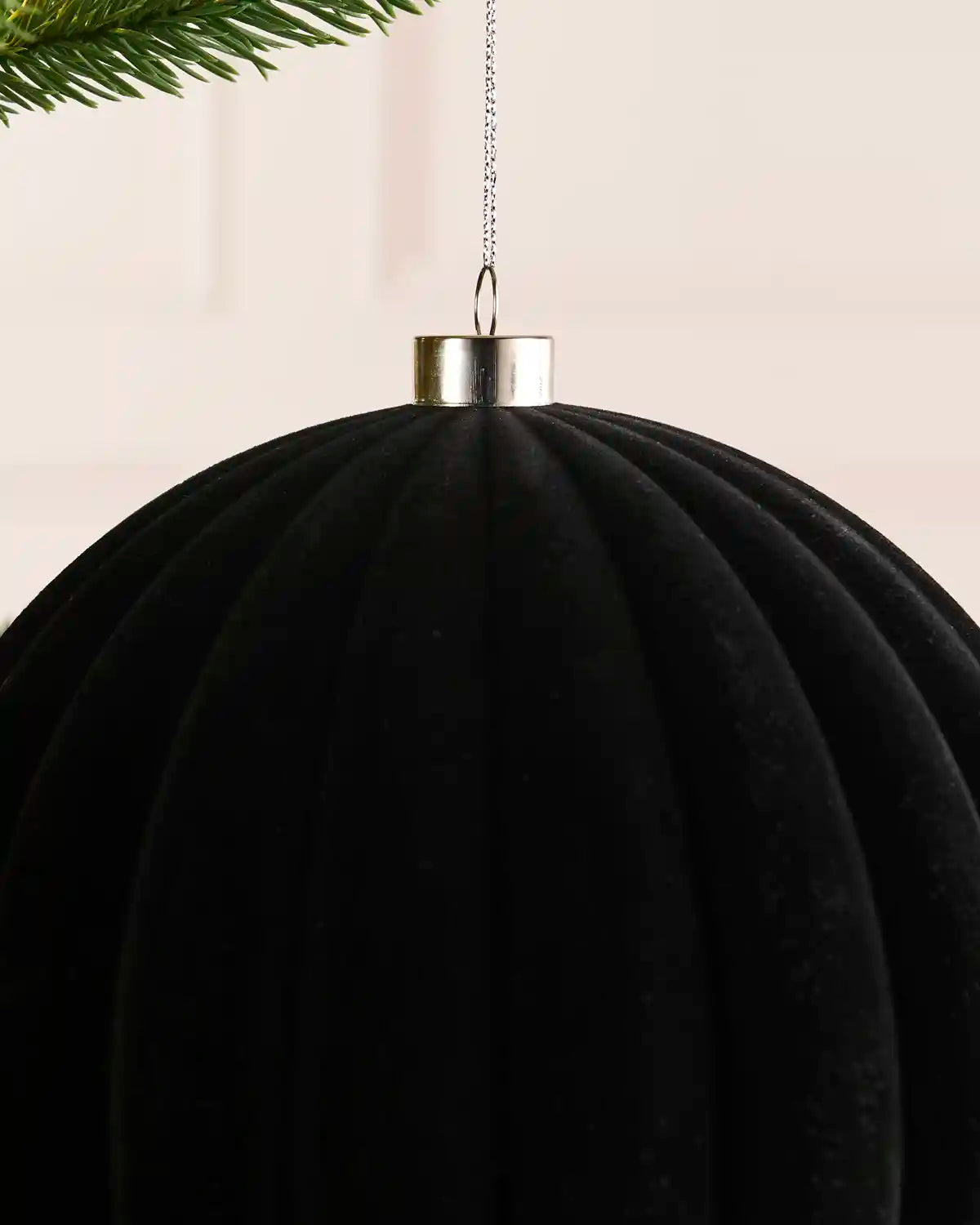 A close-up of a black ribbed hanging ornament or decorative sphere with a metallic silver cap, suspended by a thin chain against a soft light background, with a small green pine branch visible at the top left.