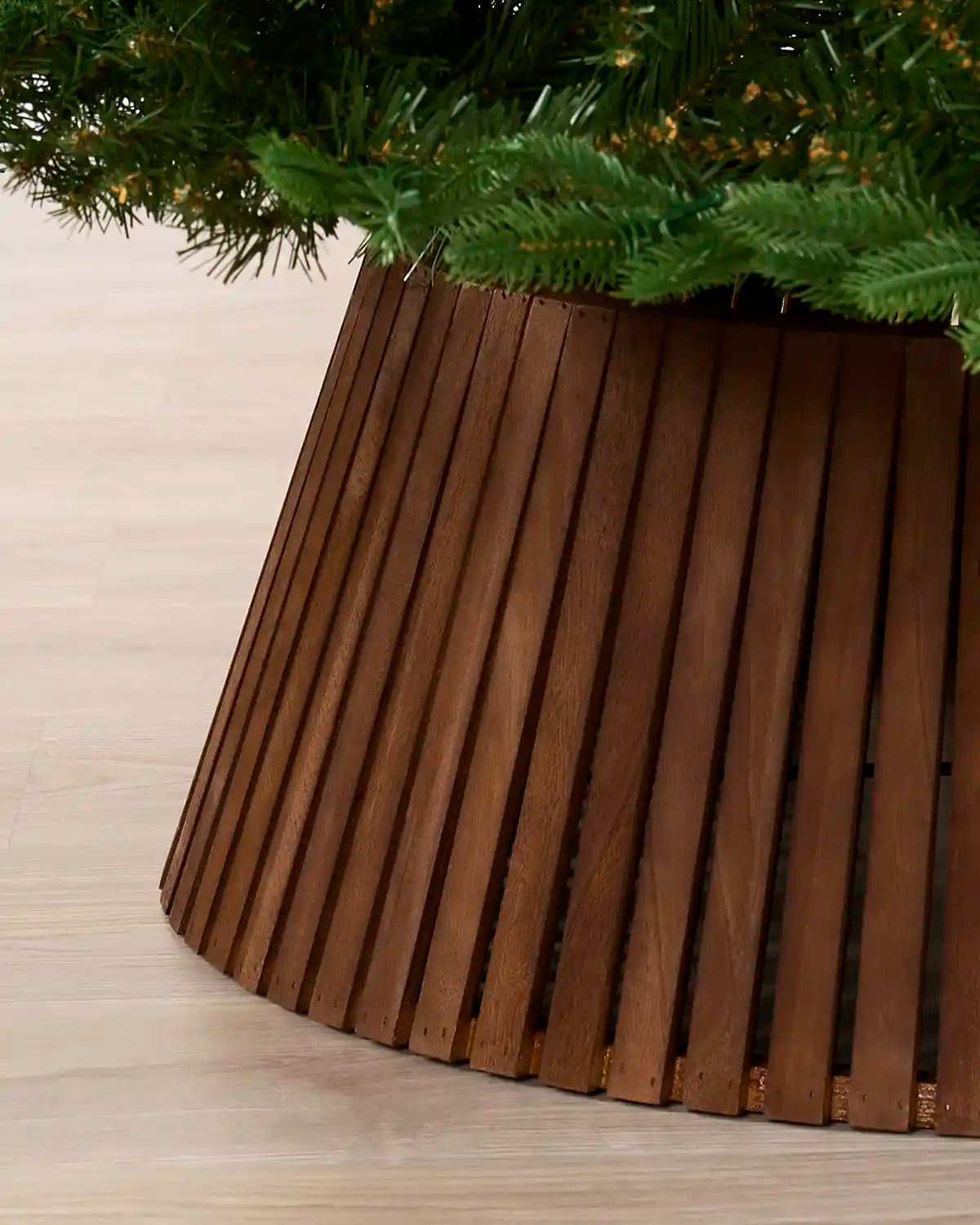 A close-up of a brown wooden slatted Christmas tree skirt supporting green artificial tree branches on a light wood floor.