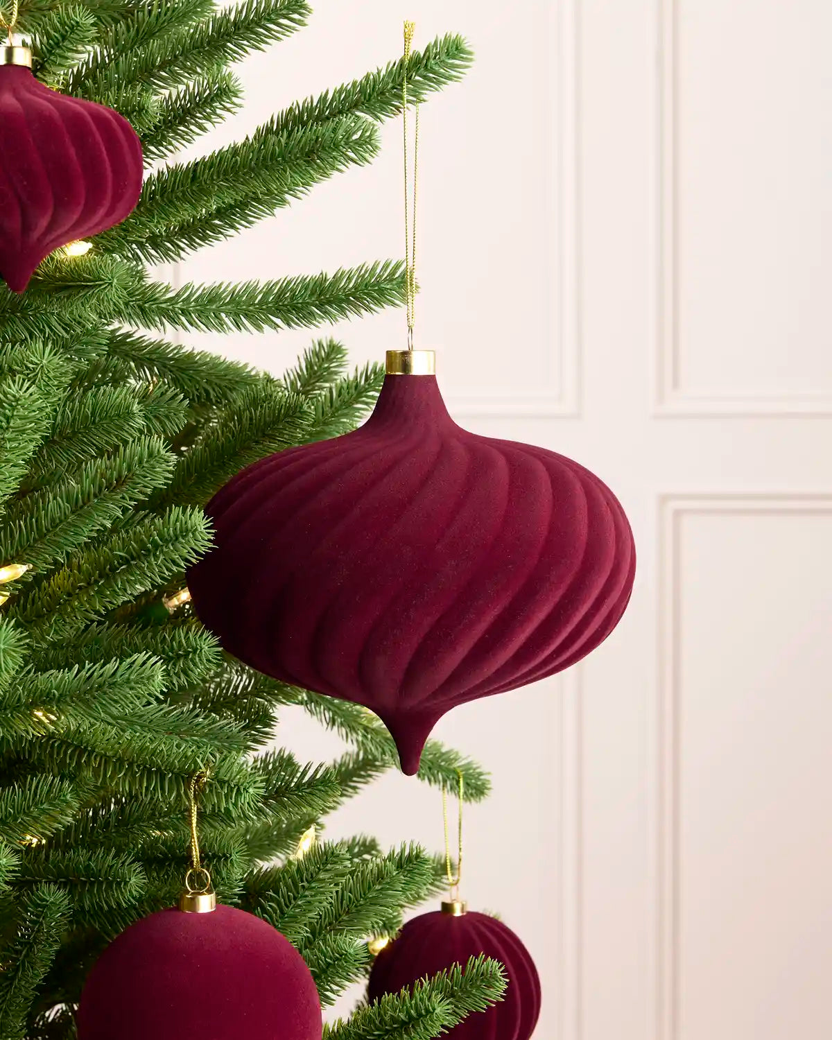 A close-up of a burgundy velvet Christmas tree ornament hanging from a green artificial Christmas tree, with a soft cream panelled wall in the background.