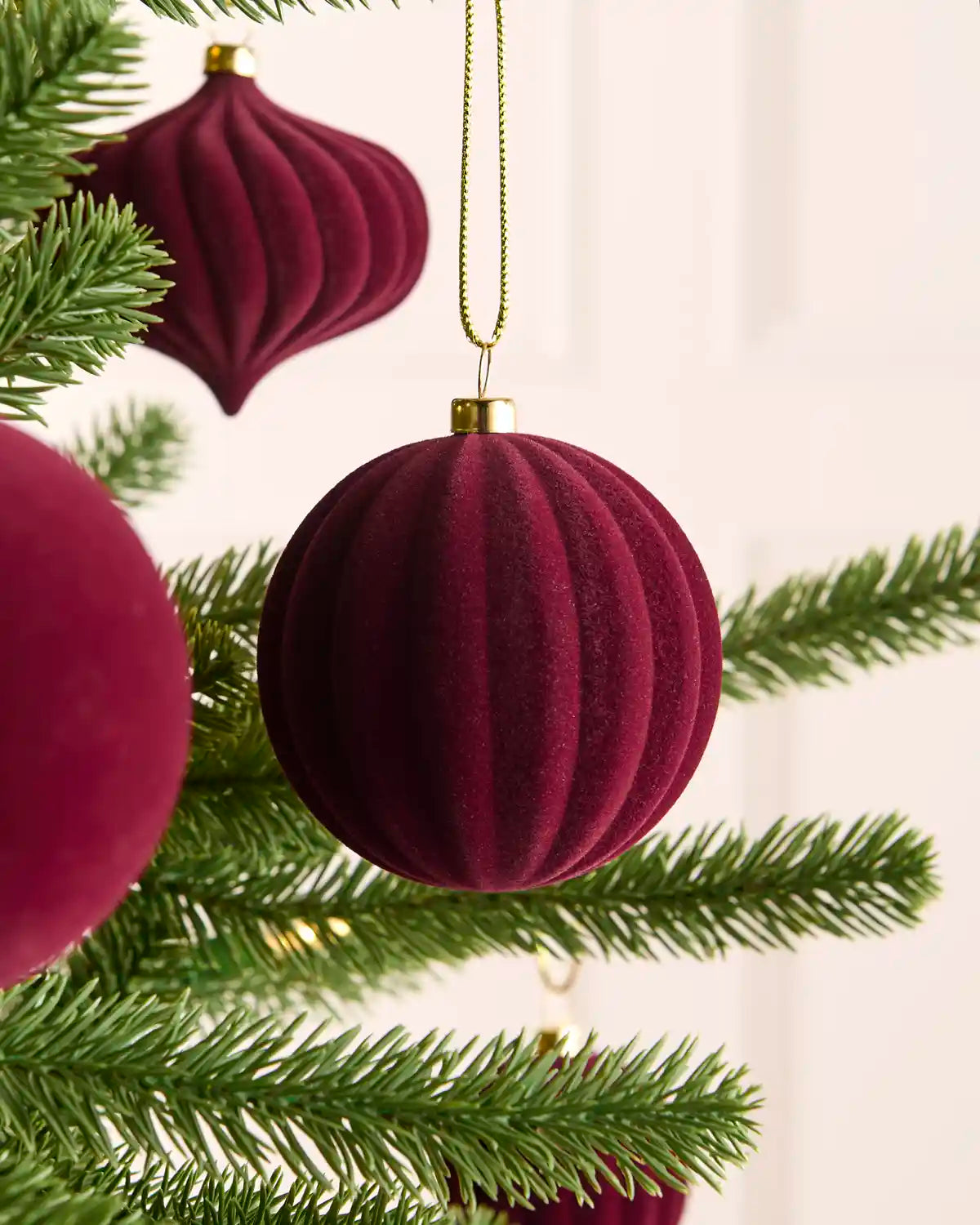 A close-up of a dark burgundy ribbed velvet Christmas ornament hanging from a gold string on a green pine tree branch, with other matching ornaments partially visible.