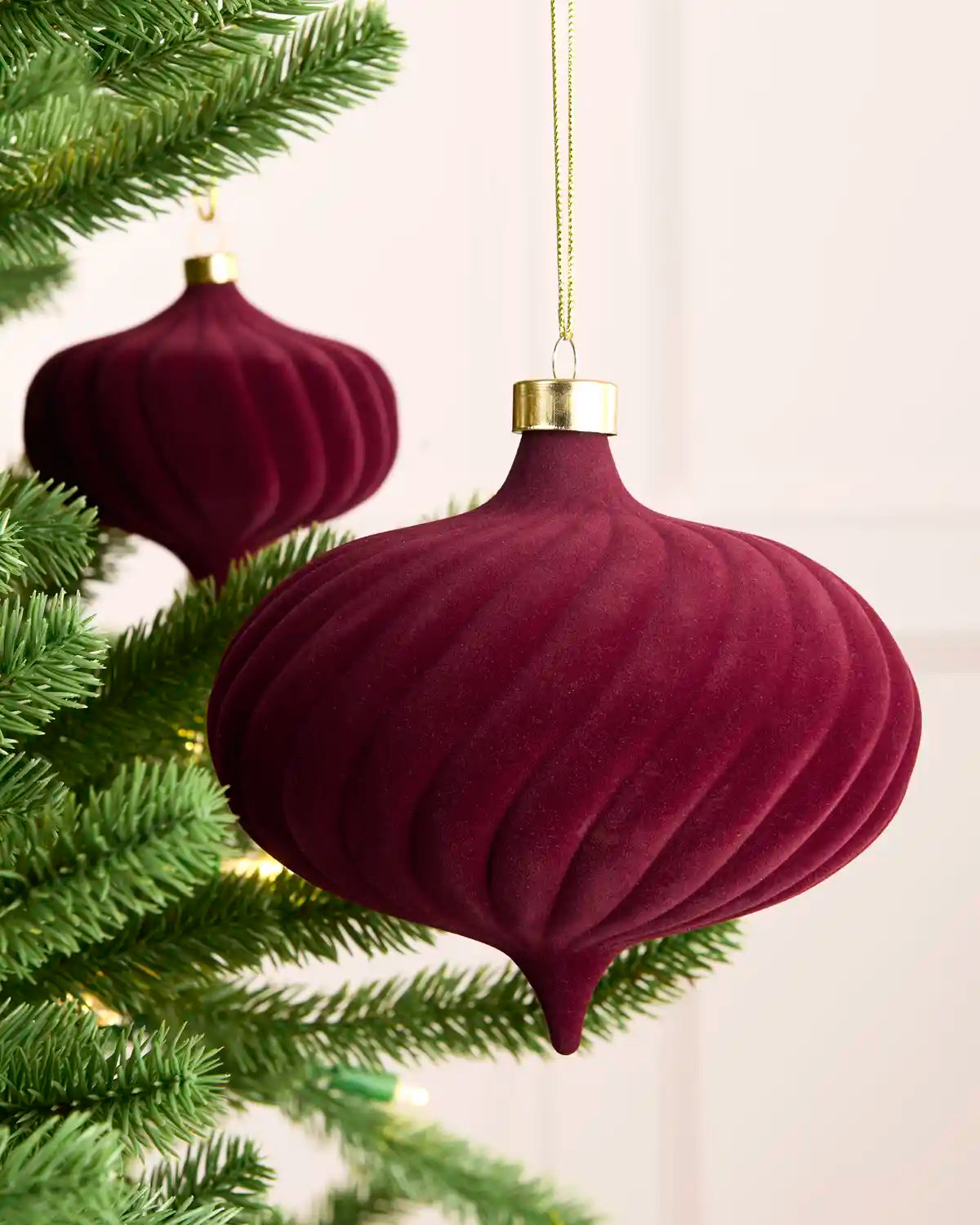 A close-up of a dark burgundy velvet Christmas ornament hanging from a green Christmas tree branch, with a second matching ornament blurred in the background.