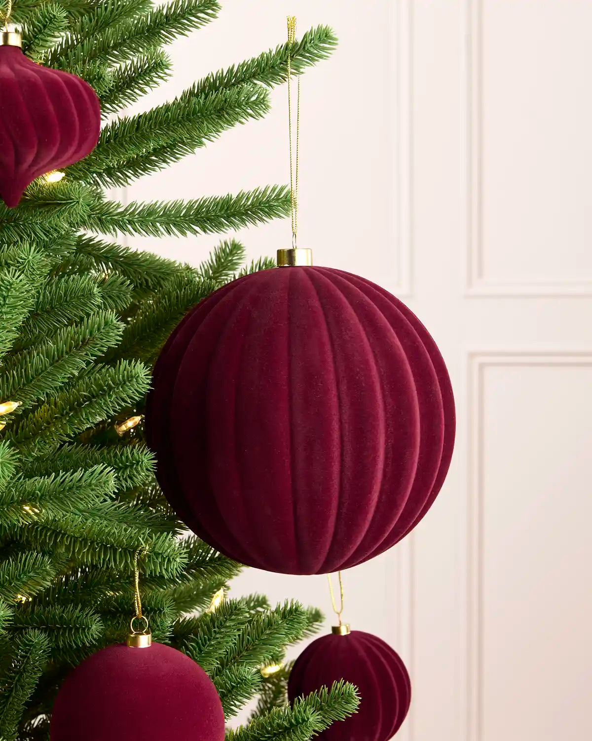 A close-up of a burgundy velvet Christmas tree ornament hanging from a green pine tree branch, with a soft cream paneled wall in the background.