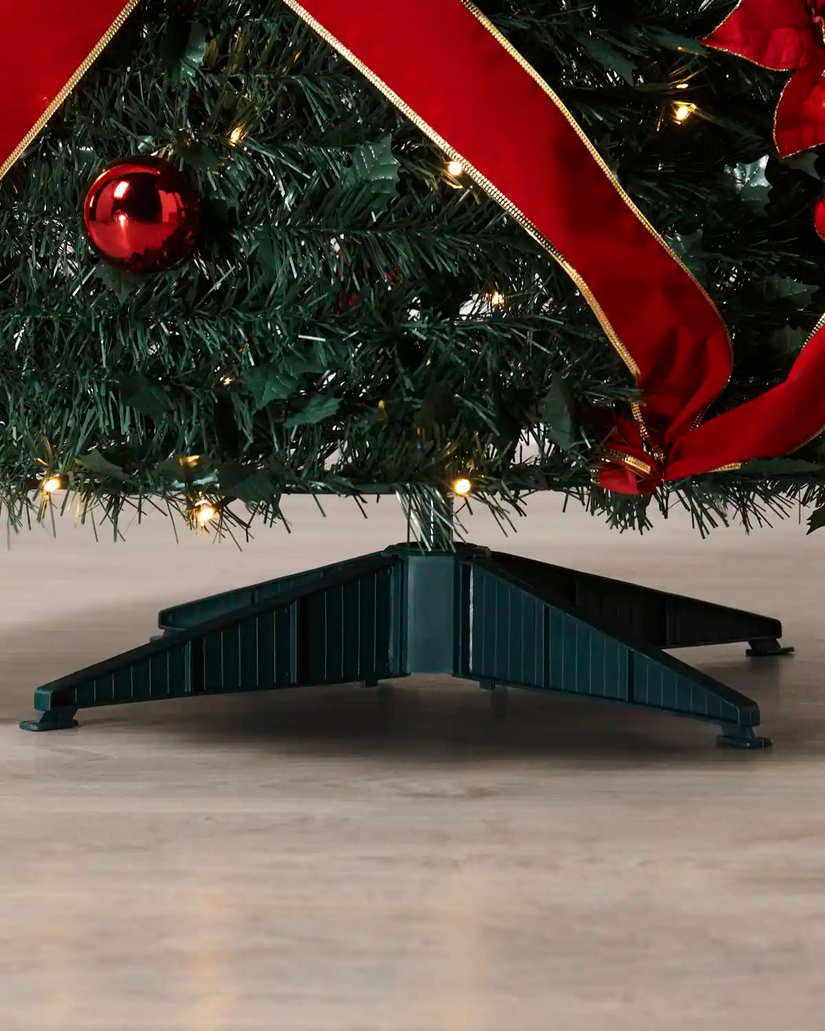 A close-up of a dark green artificial Christmas tree stand supporting a decorated tree with a red ribbon, red ornament, and warm white string lights on a light floor.