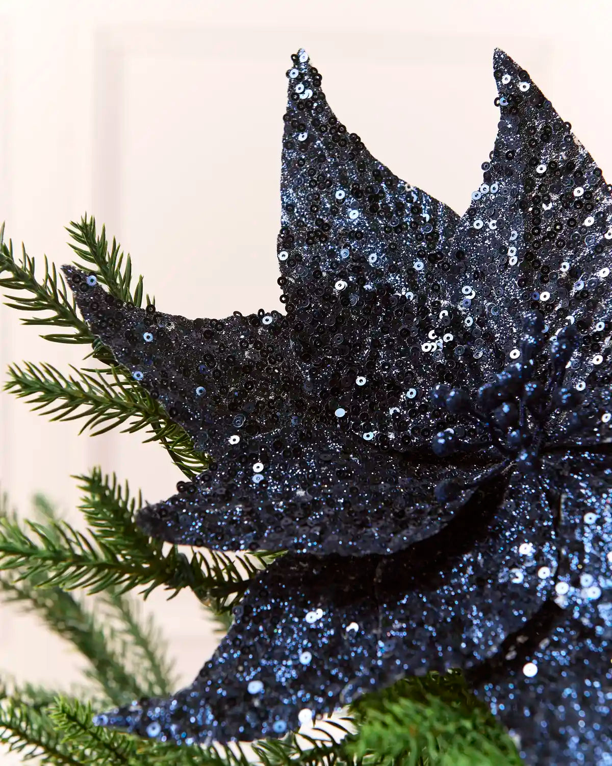 A close-up of a dark navy blue sequined poinsettia decoration resting on green pine branches against a soft, light beige background.