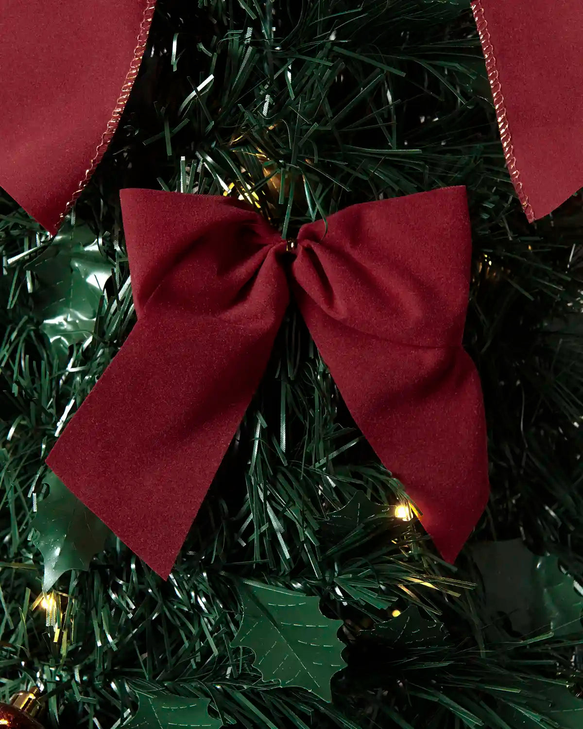 A close-up of a dark red fabric bow tied around a green artificial Christmas tree with small warm lights and holiday decorations.