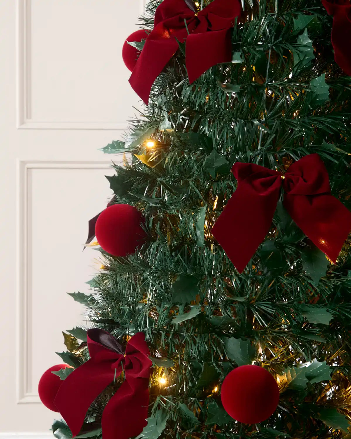 A close-up of a decorated Christmas tree with Holly leaves, dark green artificial branches, shiny dark red ornaments, large dark red ribbon bows, and warm white string lights