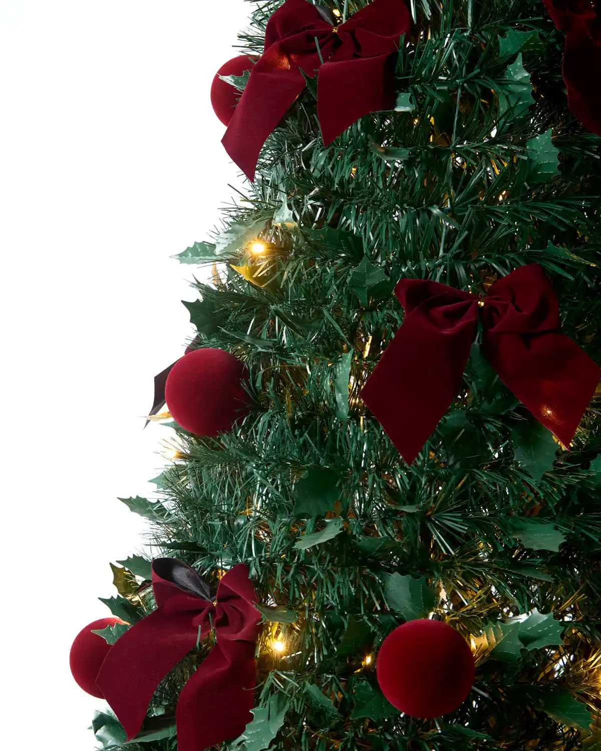 A close-up of a decorated Christmas tree with Holly leaves, dark green artificial branches, shiny dark red ornaments, large dark red ribbon bows, and warm white string lights