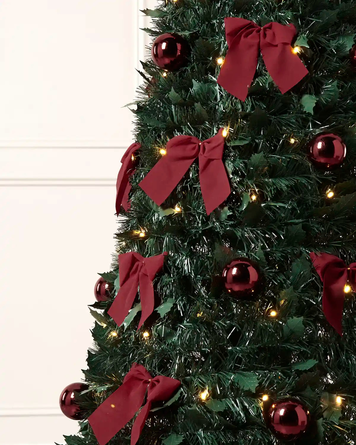 A close-up of a decorated Christmas tree with Holly leaves, dark green artificial branches, shiny dark red ornaments, large dark red ribbon bows, and warm white string lights against an off-white panelled wall.