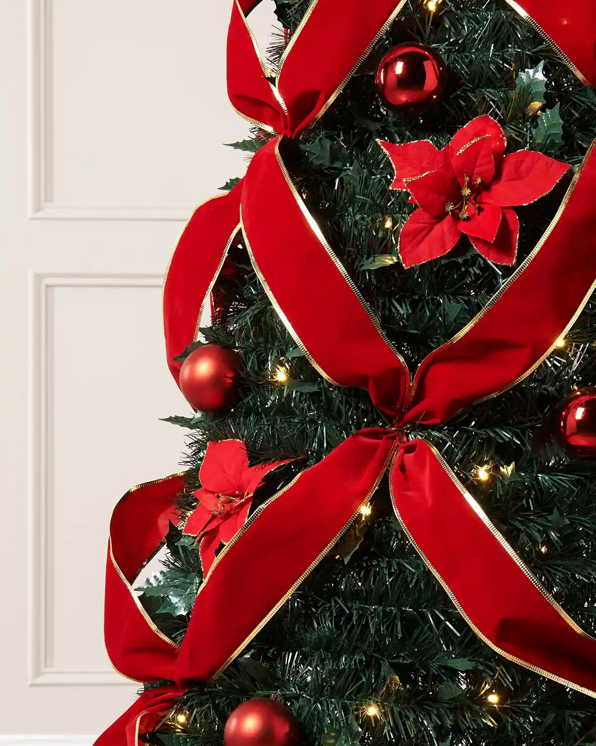 A close-up of a decorated Christmas tree with a large red ribbon bow, red poinsettia ornaments, red baubles, and warm white string lights against a white panneled door background.