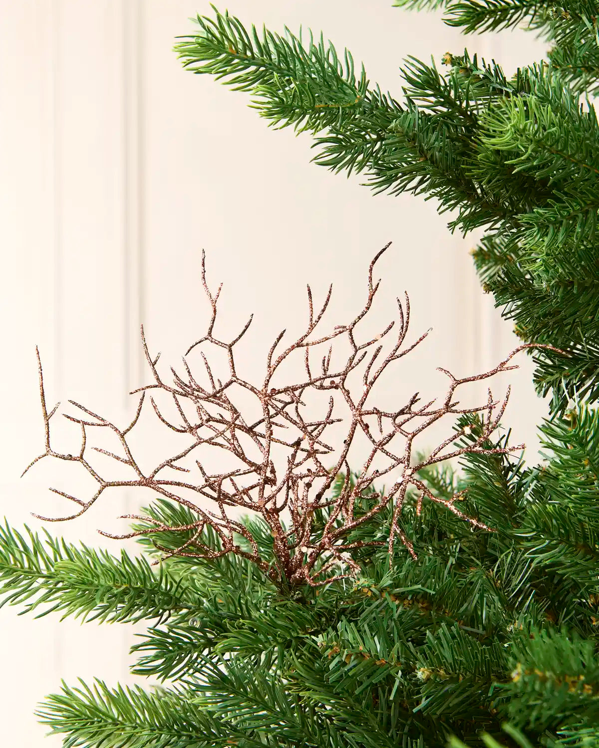 A close-up of a decorative Espresso  branch arrangement placed among the green needles of a Christmas tree, with a soft cream-colored panelled wall in the background.