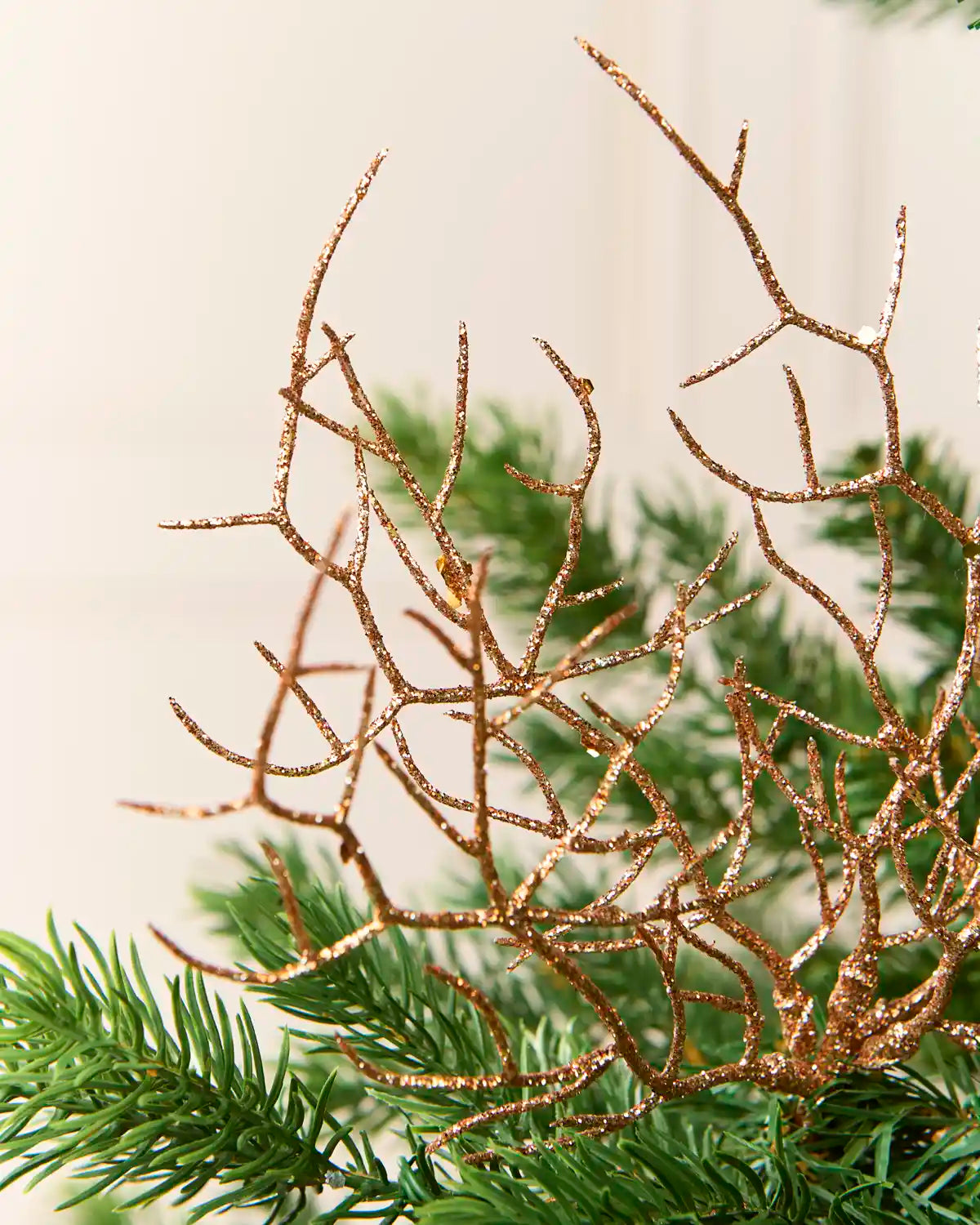 A close-up of a decorative Rose-gold glitter branch ornament with a textured, twig-like structure, shown against a soft, out-of-focus background of green Christmas tree needles and a light cream wall.