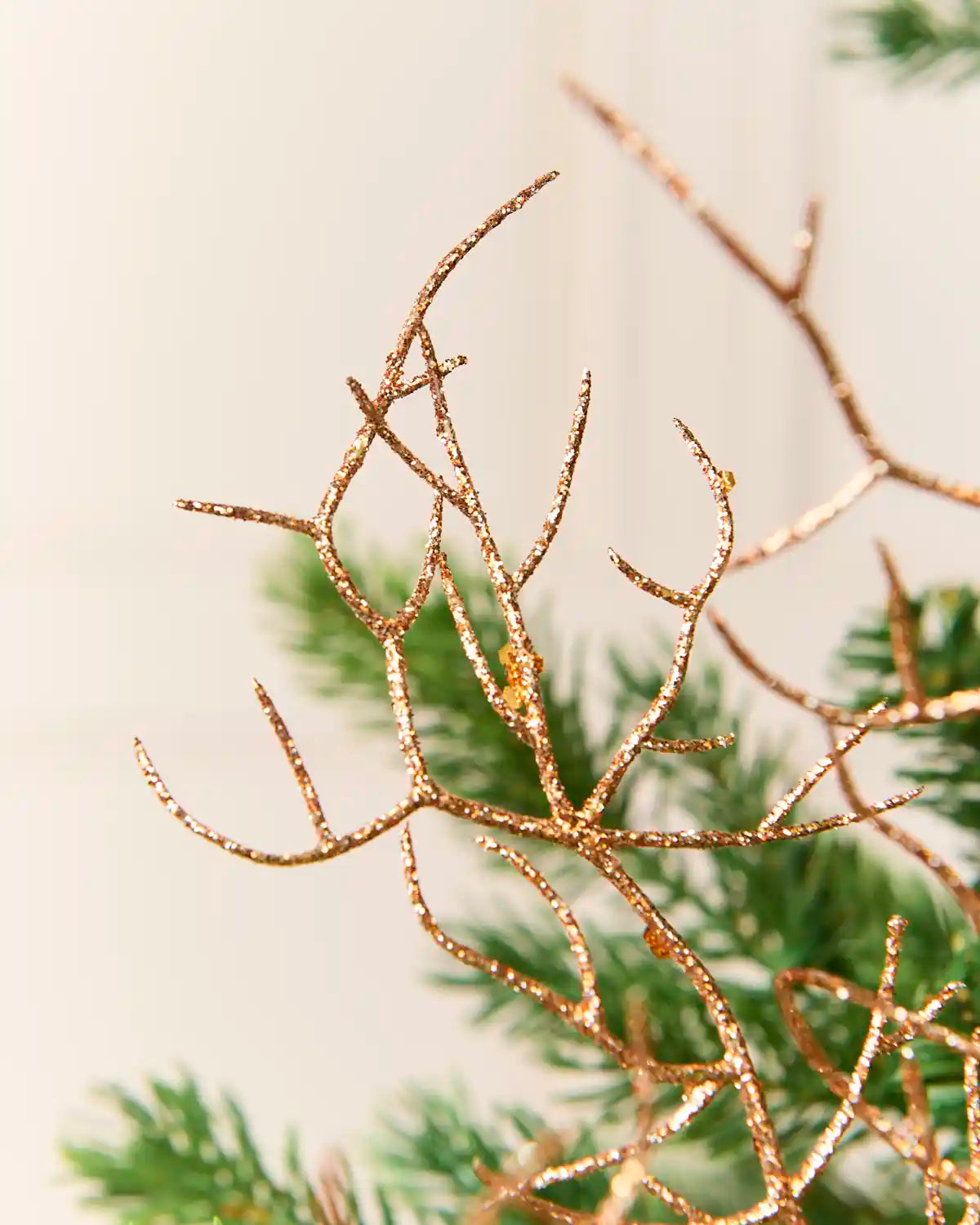 A close-up of a decorative Rose-gold glitter branch ornament with a textured, twig-like structure, shown against a soft, out-of-focus background of green pine needles and a light cream wall.