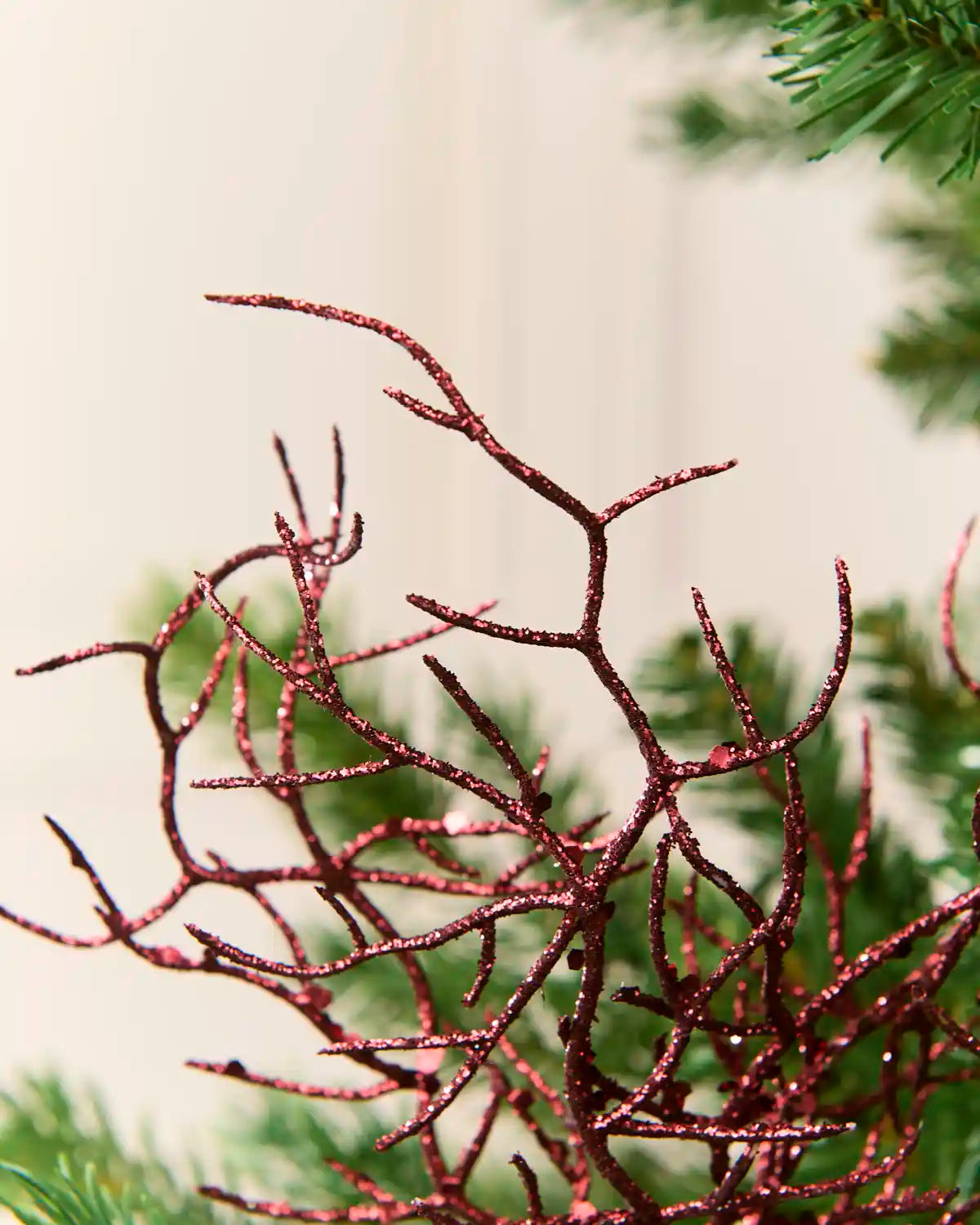 A close-up of a decorative coral ornament with a glittery burgundy red finish, styled among green pine needles against a soft cream background.