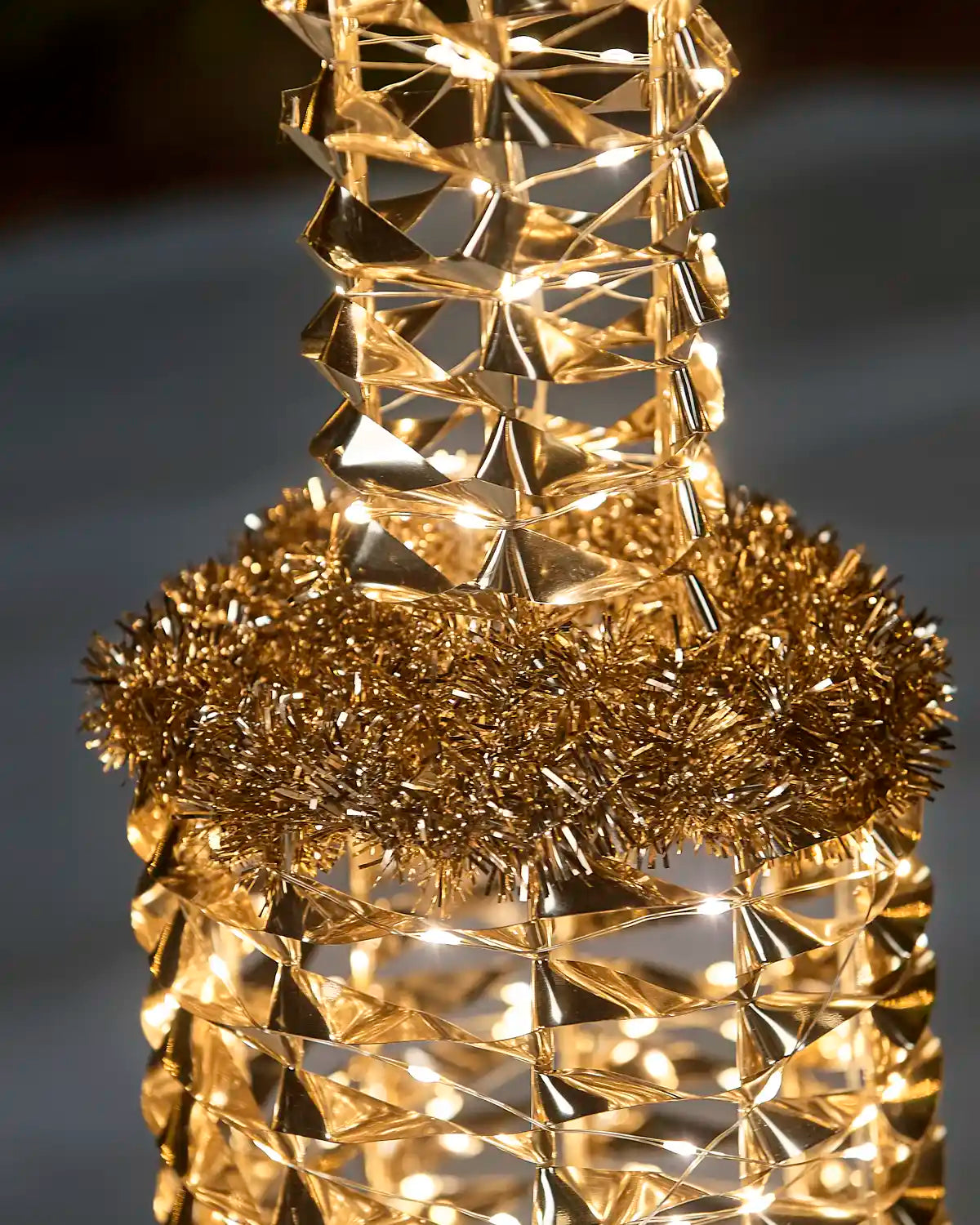 A close-up of a decorative gold and warm white LED lamppost with a fluffy gold tinsel collar, glowing with warm lights against a dark blurred background.