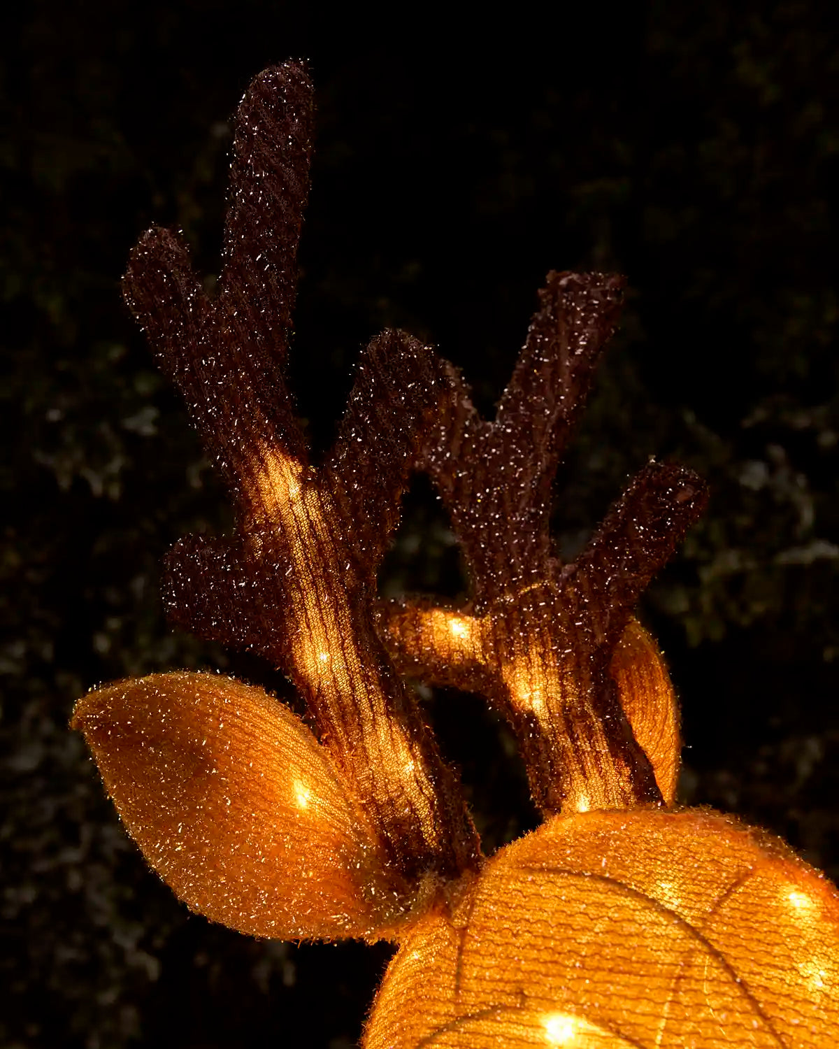 A close-up of a decorative golden and brown reindeer head light or ornament, with glowing warm lights visible through a textured netted surface against a dark outdoor background.