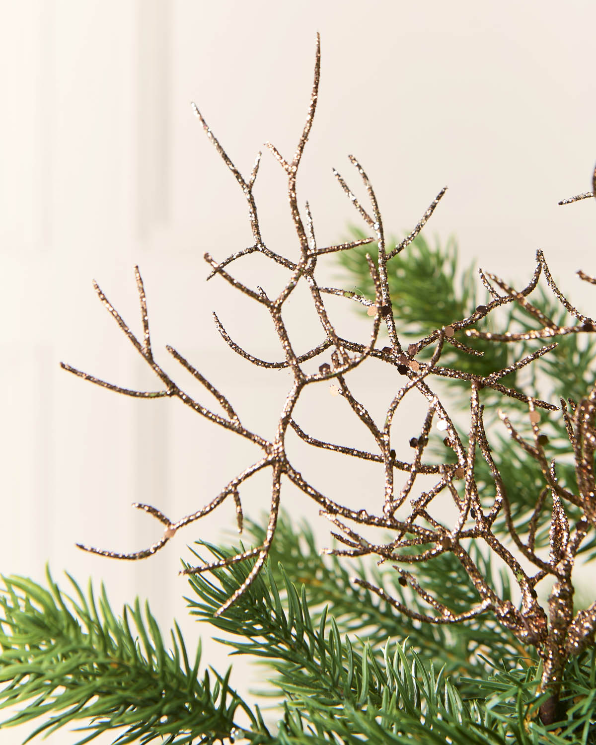A close-up of a decorative tree branch ornament with glittery Mocha branches layered over green pine needles, set against a soft cream background.