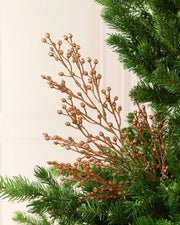 A close-up of a decorative tree branch with metallic Rose gold berries and small cone-like buds, layered over green pine needles against a soft off-white panelled background.