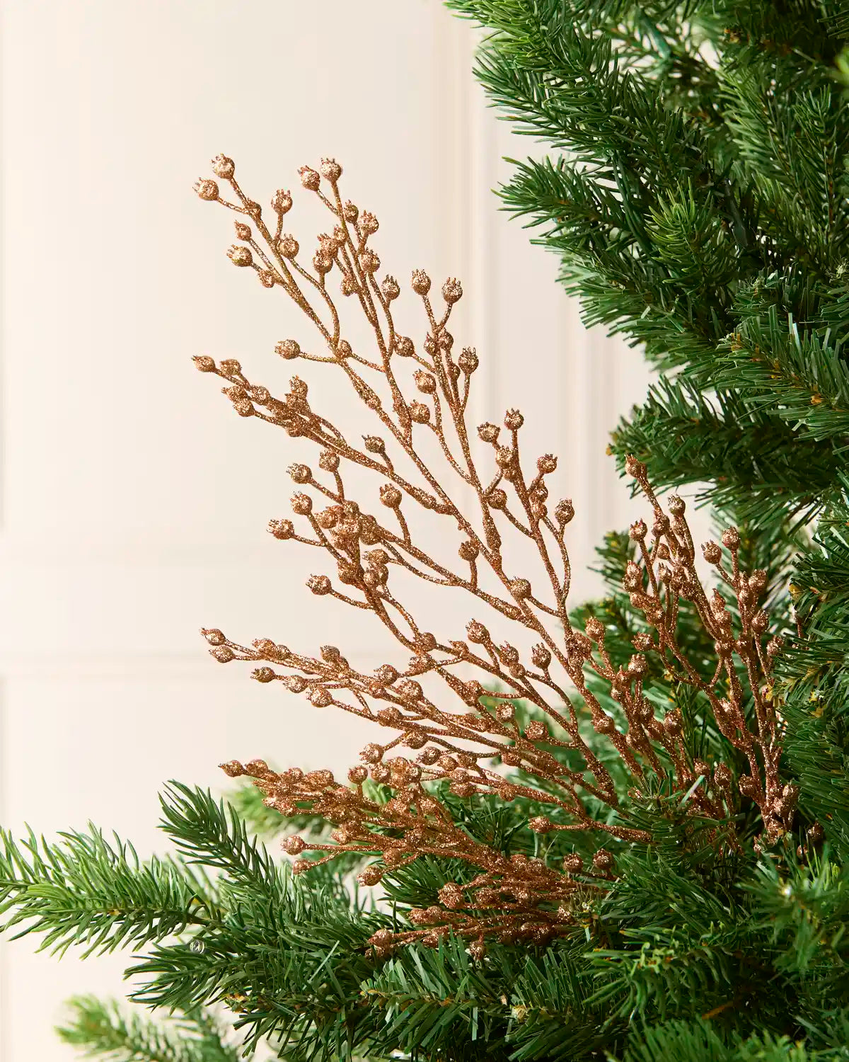 A close-up of a decorative tree branch with metallic Rose gold berries and small cone-like buds, layered over green pine needles against a soft off-white panelled background.