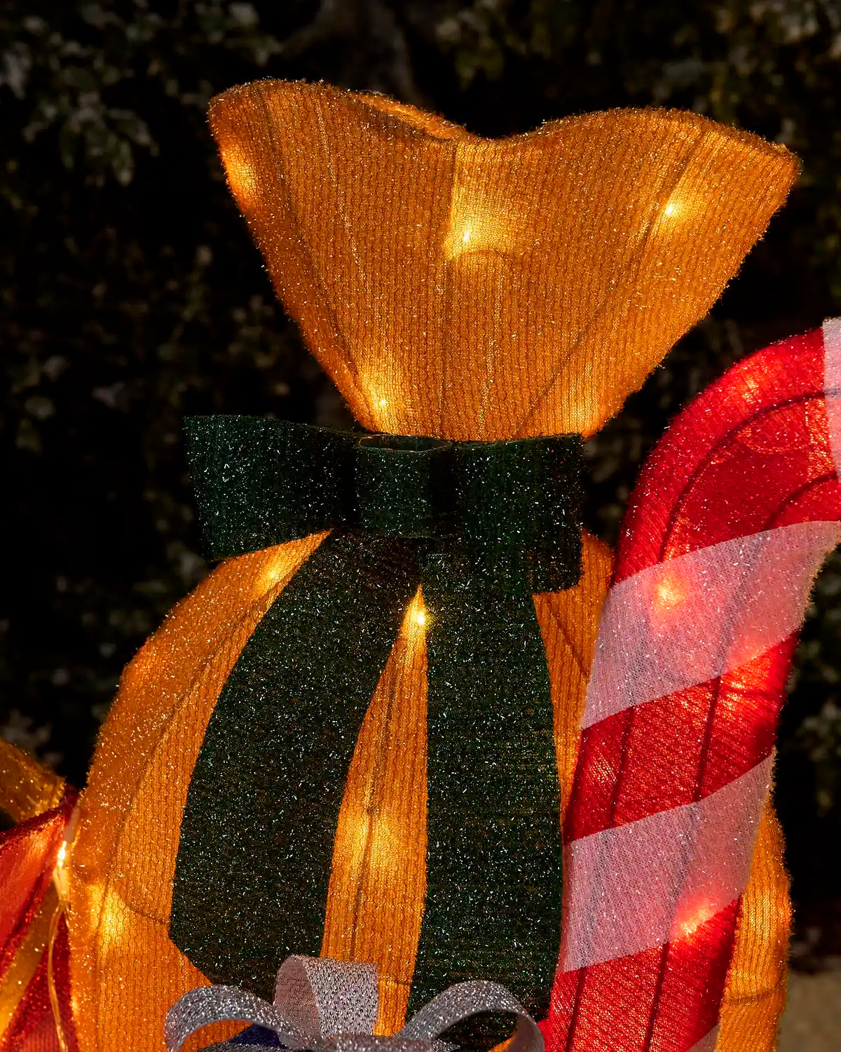 A close-up of a festive holiday decoration featuring a glowing gold gift bag with a dark green glittery bow, warm white string lights, and part of a red-and-white striped candy cane visible on the right.