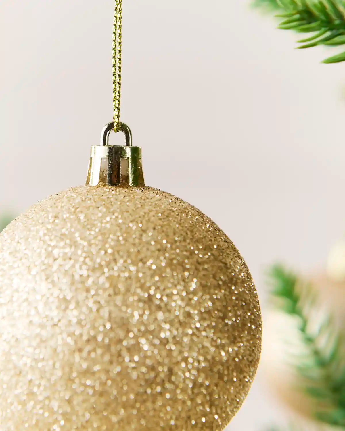 A close-up of a glittery Latte Christmas ornament hanging from a gold chain, with soft blurred green pine branches in the background.