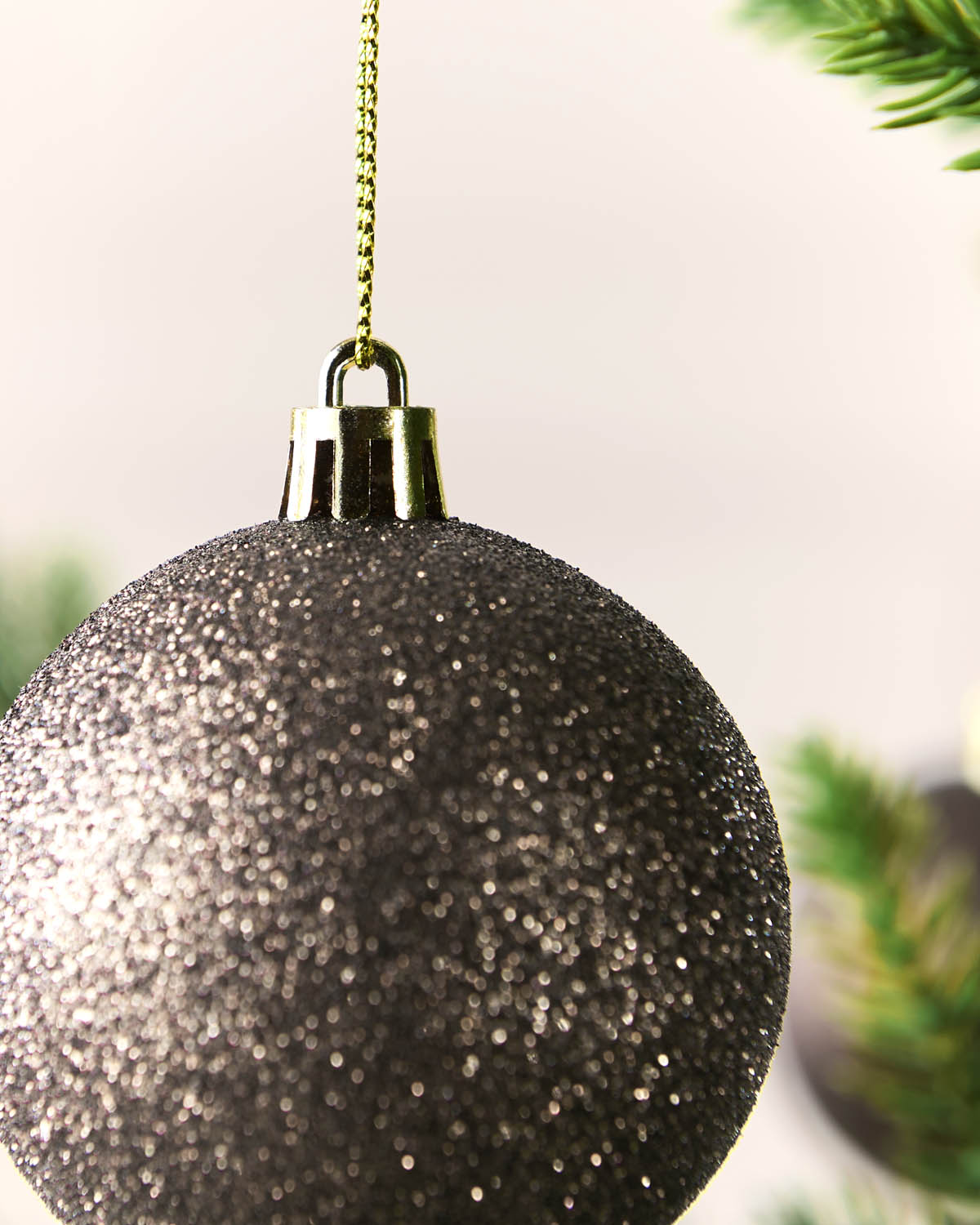 A close-up of a glittery Mocha Christmas ornament hanging from a gold chain, with blurred green pine branches in the background.