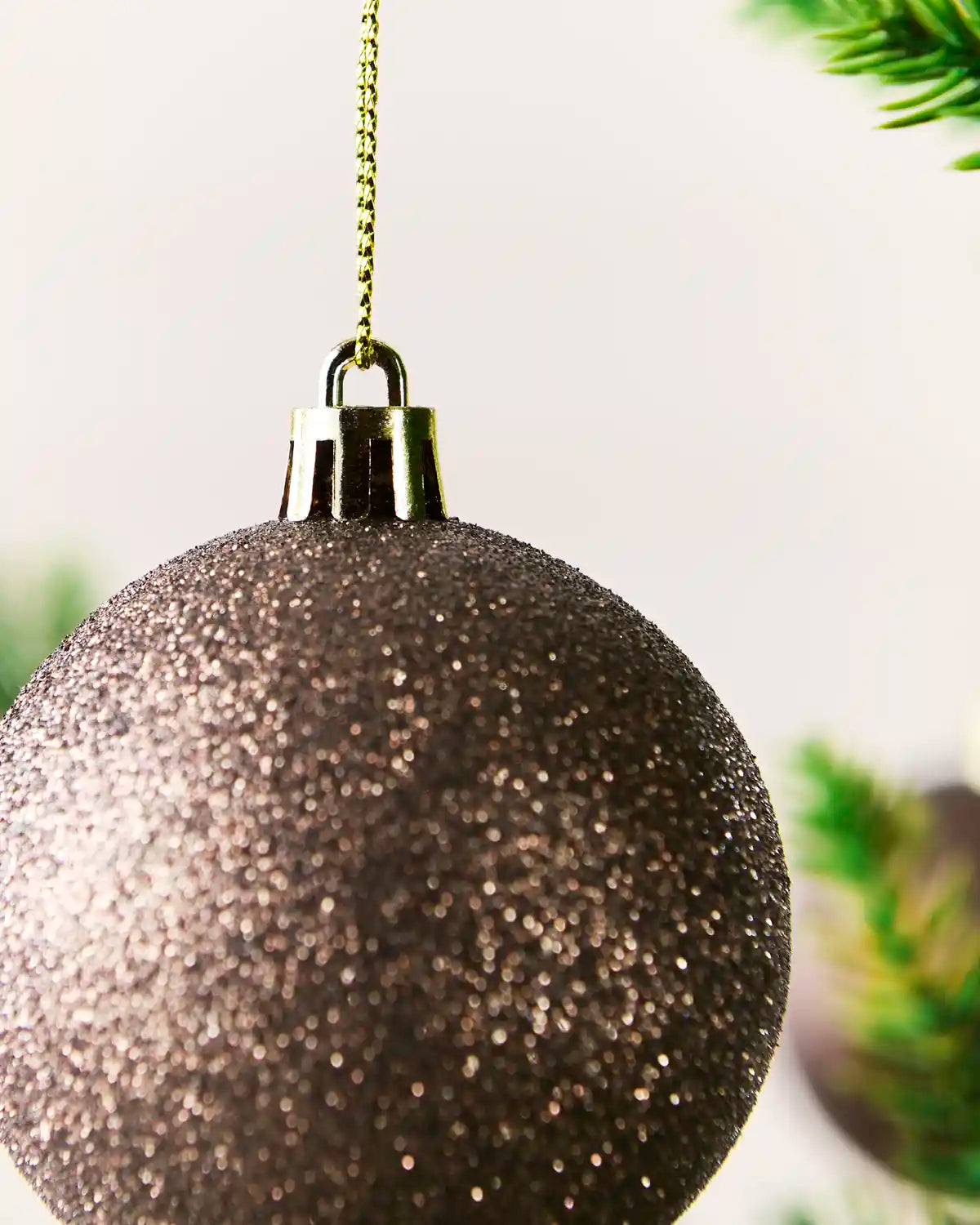 A close-up of a glittery Mocha Christmas ornament hanging from a gold chain, with blurred green pine branches in the background.