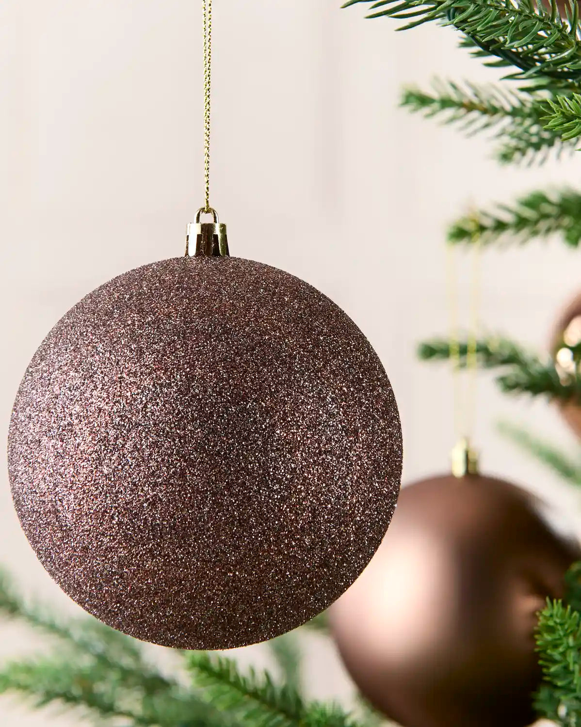 A close-up of a glittery Mocha Christmas tree ornament hanging from a gold string, with blurred pine branches and another metallic ornament in the background.