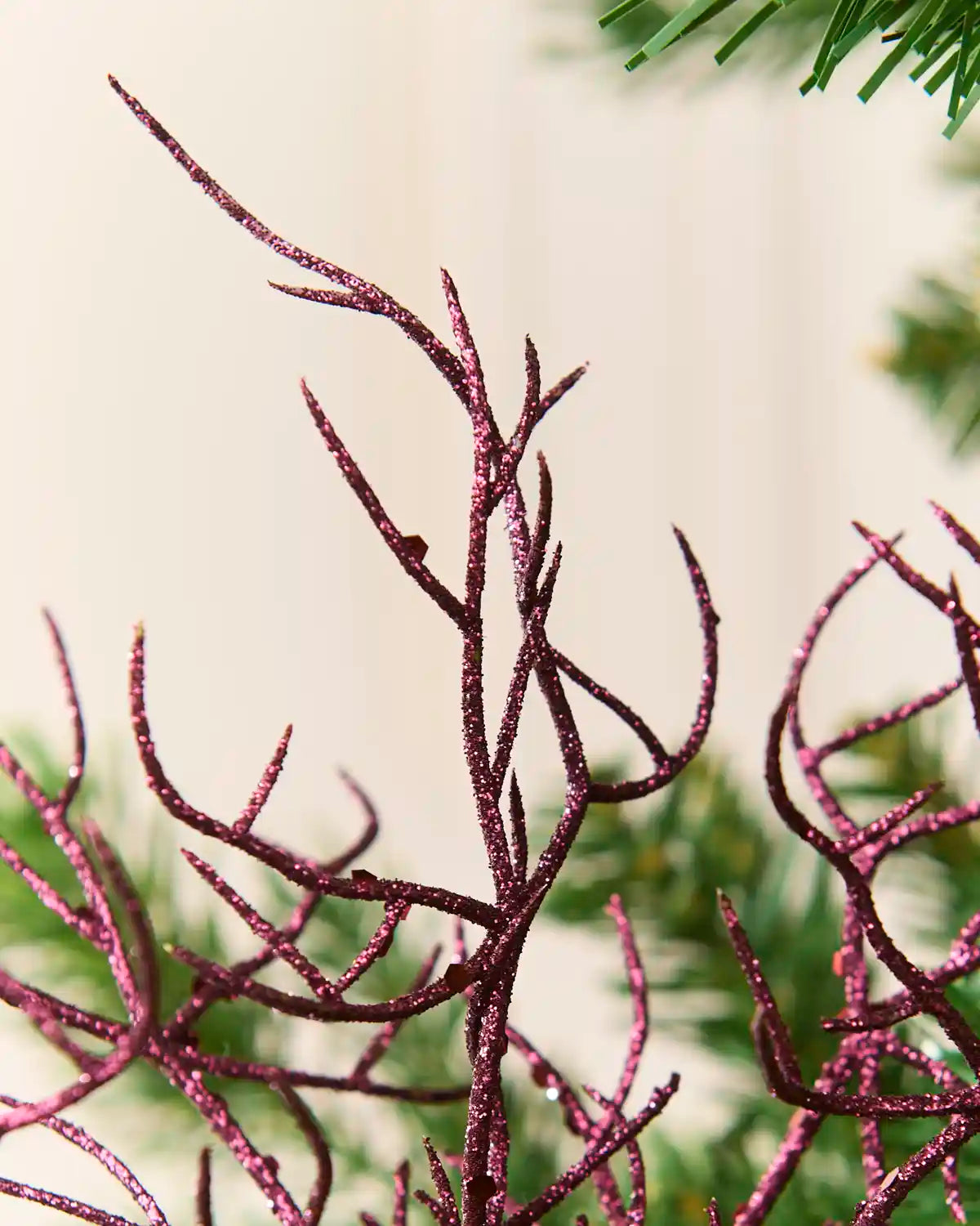 A close-up of a glittery Purple decorative branch or coral-style ornament attached to green artificial Christmas tree branches against a soft cream background. 