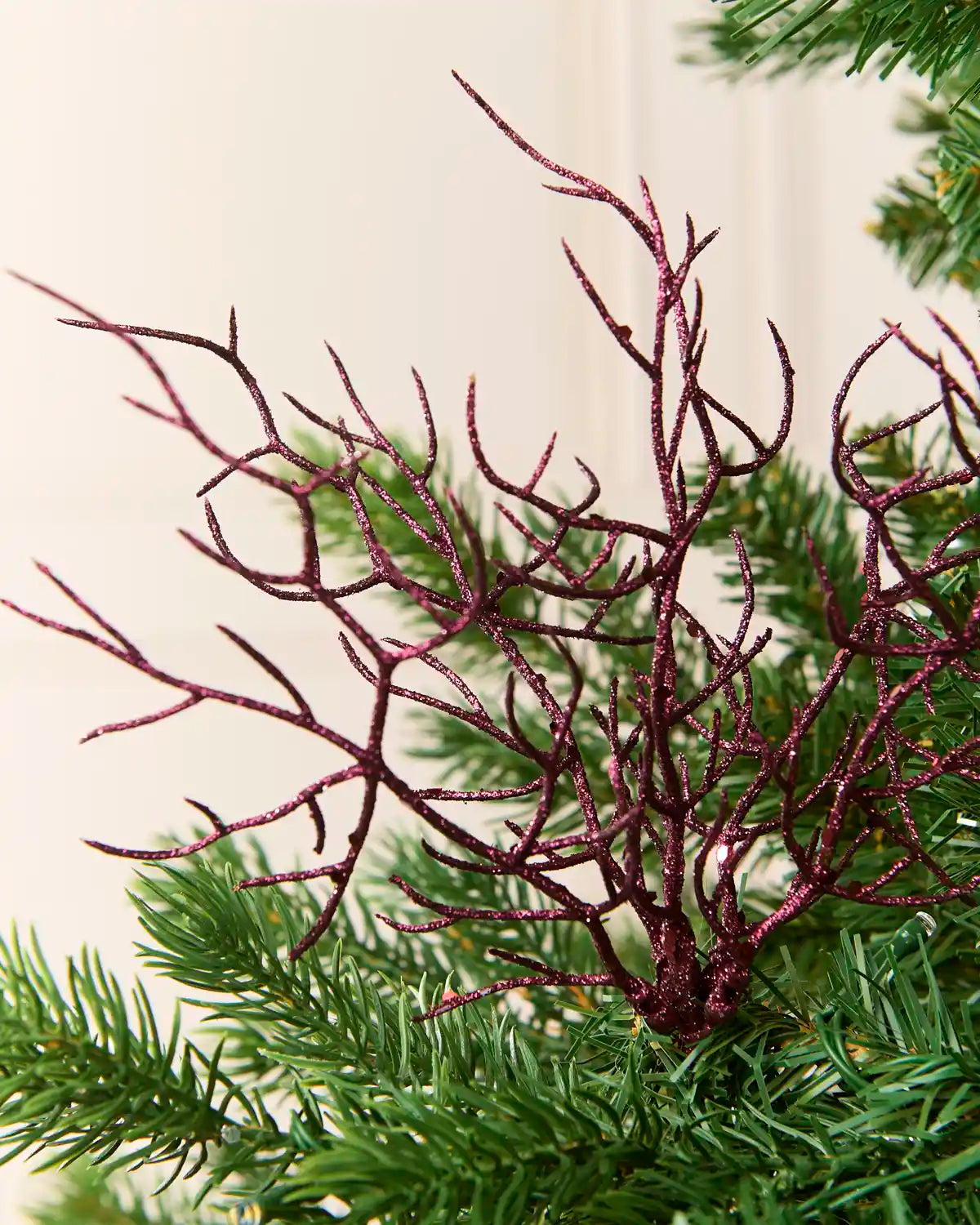 A close-up of a glittery Purple decorative branch or coral-style ornament attached to green artificial pine branches against a soft cream background.