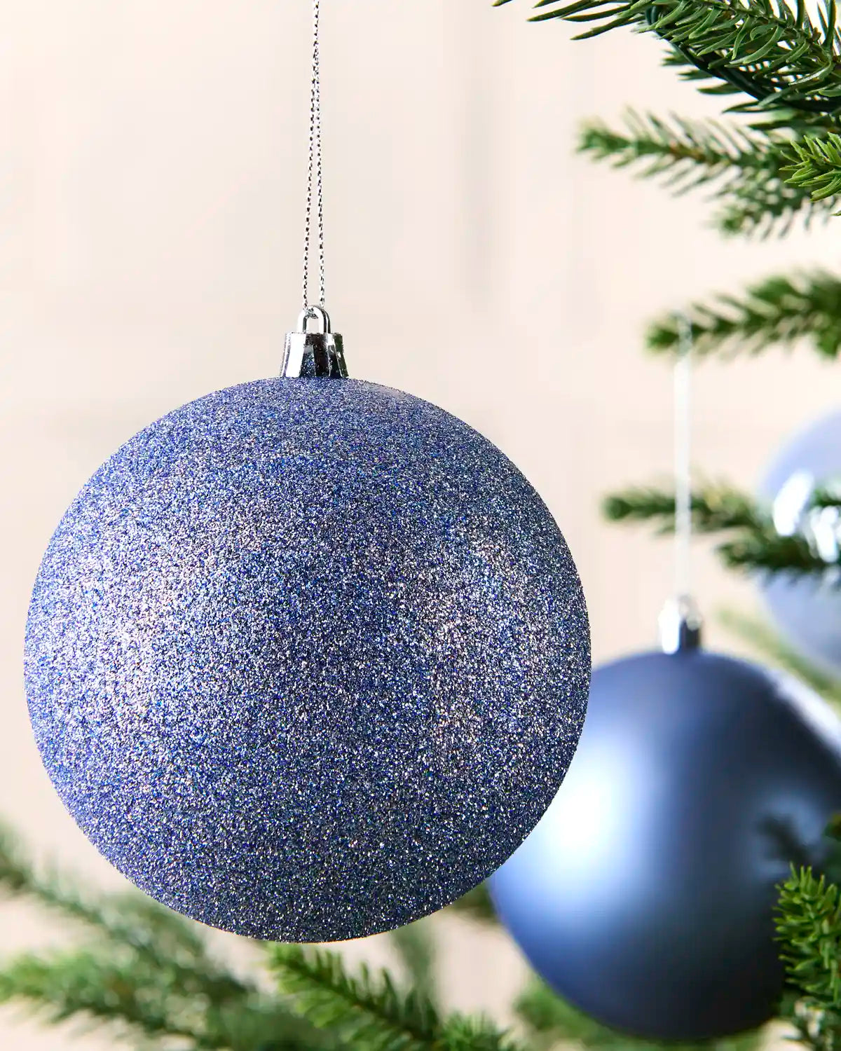 A close-up of a glittery blue Christmas ornament hanging from a silver cap on a tree branch, with other blue ornaments and green pine needles softly blurred in the background.