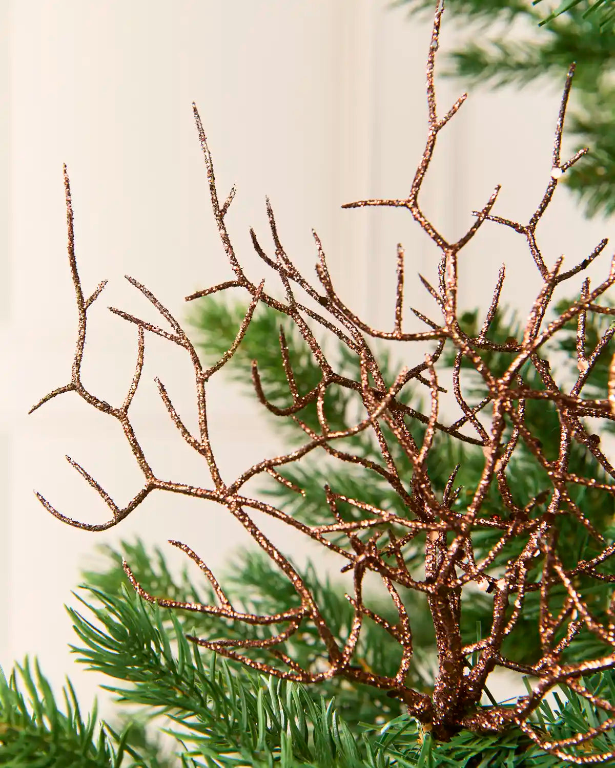 A close-up of a glittery espresso decorative branch or tree topper with fine twig-like limbs, shown against a soft cream background with green pine needles in the lower right.