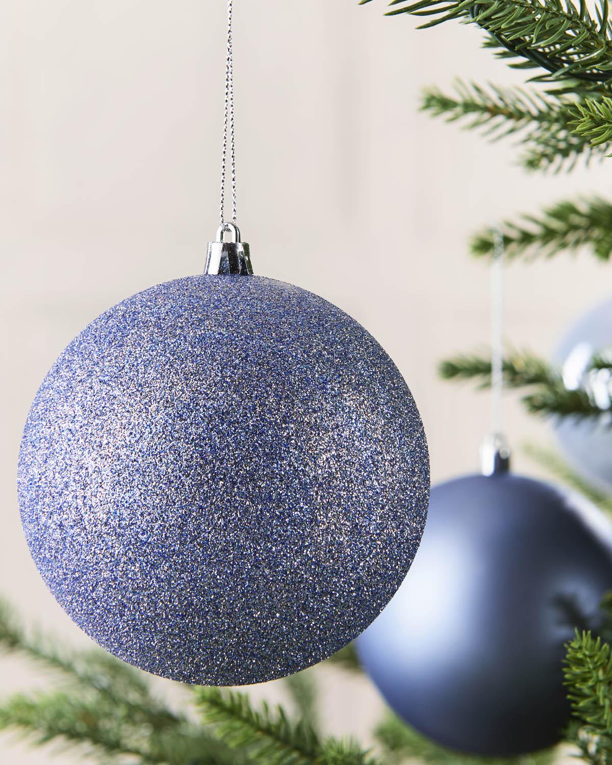 A close-up of a glittery ice blue Christmas ornament hanging from a tree branch, with other blurred ornaments and pine needles in the background.