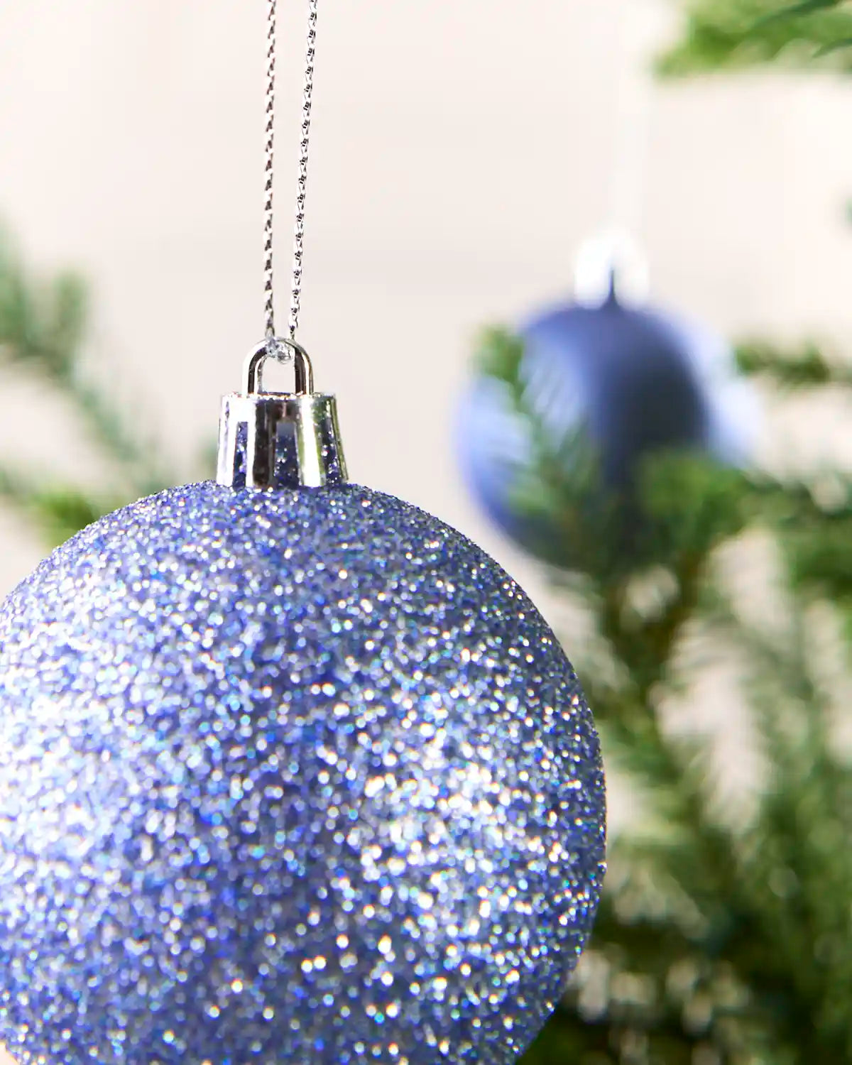 A close-up of a glittery light blue Christmas ornament hanging from a silver cap on a silver chain, with a blurred second blue ornament and green tree branches in the background.