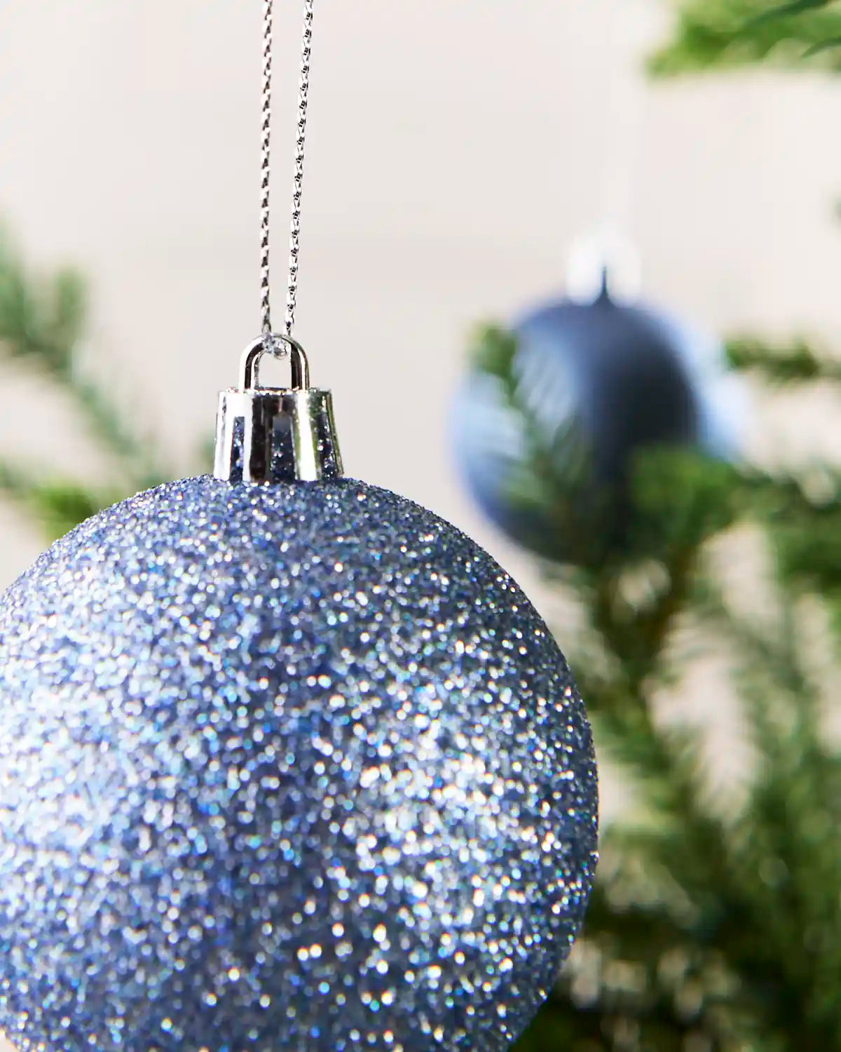A close-up of a glittery light blue Christmas ornament hanging from a silver chain, with blurred green tree branches and another ornament in the background.