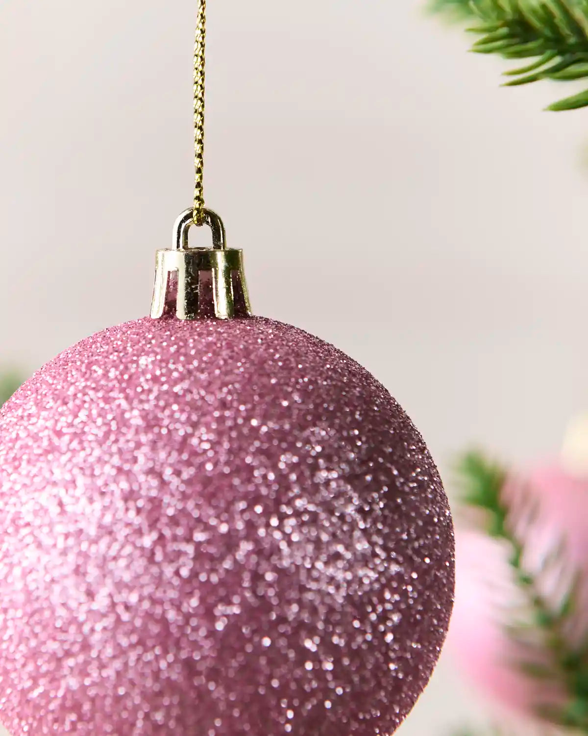 A close-up of a glittery pink Christmas ornament hanging from a gold chain, with blurred green pine branches in the background.