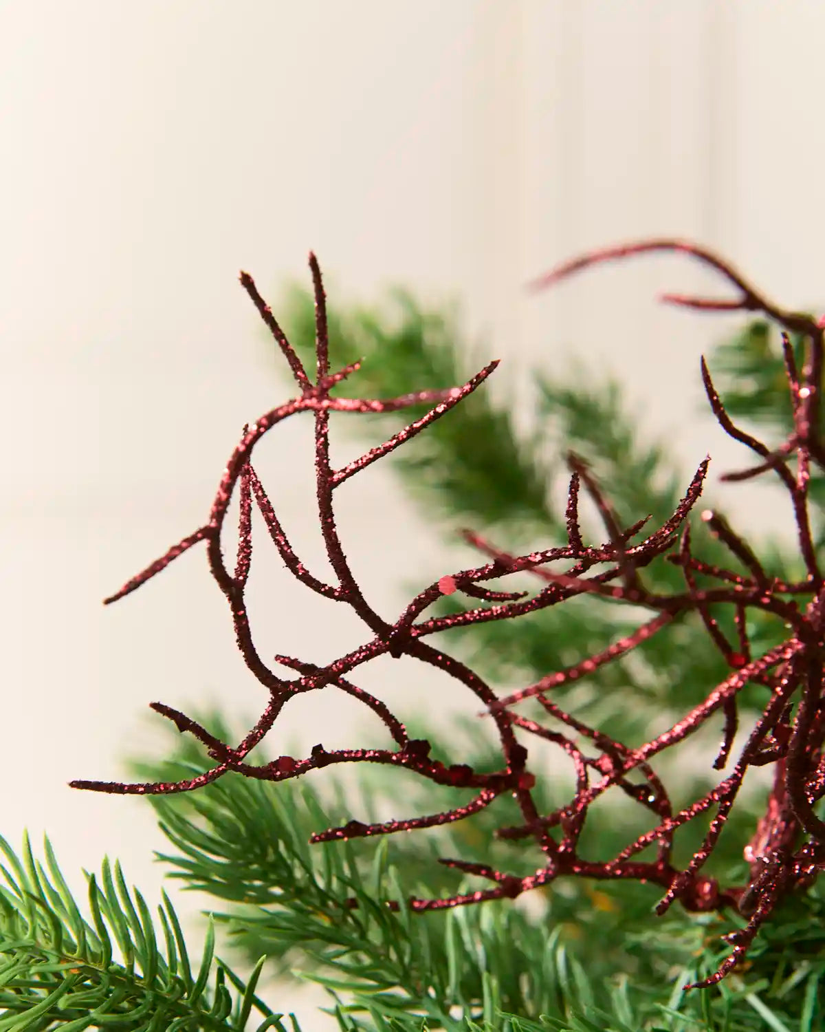 A close-up of a glittery red decorative branch or pine branch ornament layered over green pine needles, with a soft cream background.