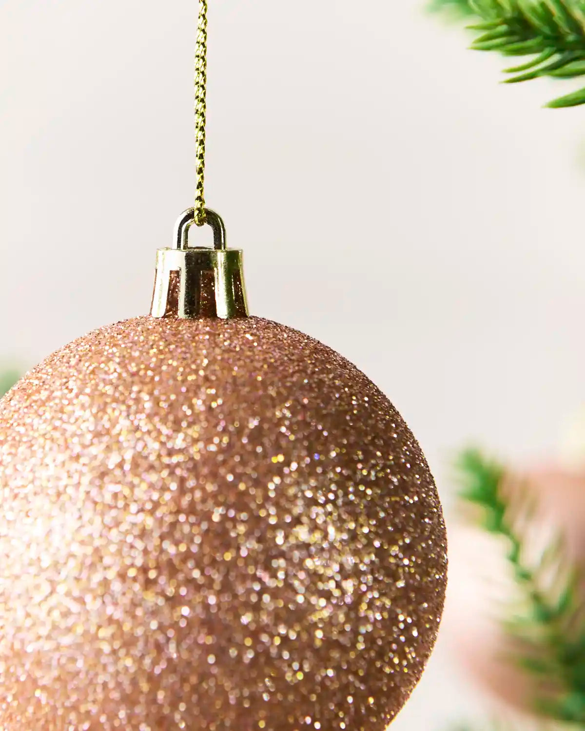 A close-up of a glittery rose gold Christmas ornament hanging from a gold chain, with soft-focus pine branches in the background.