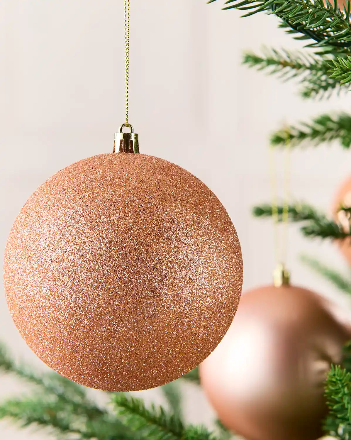 A close-up of a glittery rose gold Christmas ornament hanging from a gold string on a Christmas tree, with other blurred ornaments and tree branches in the background.