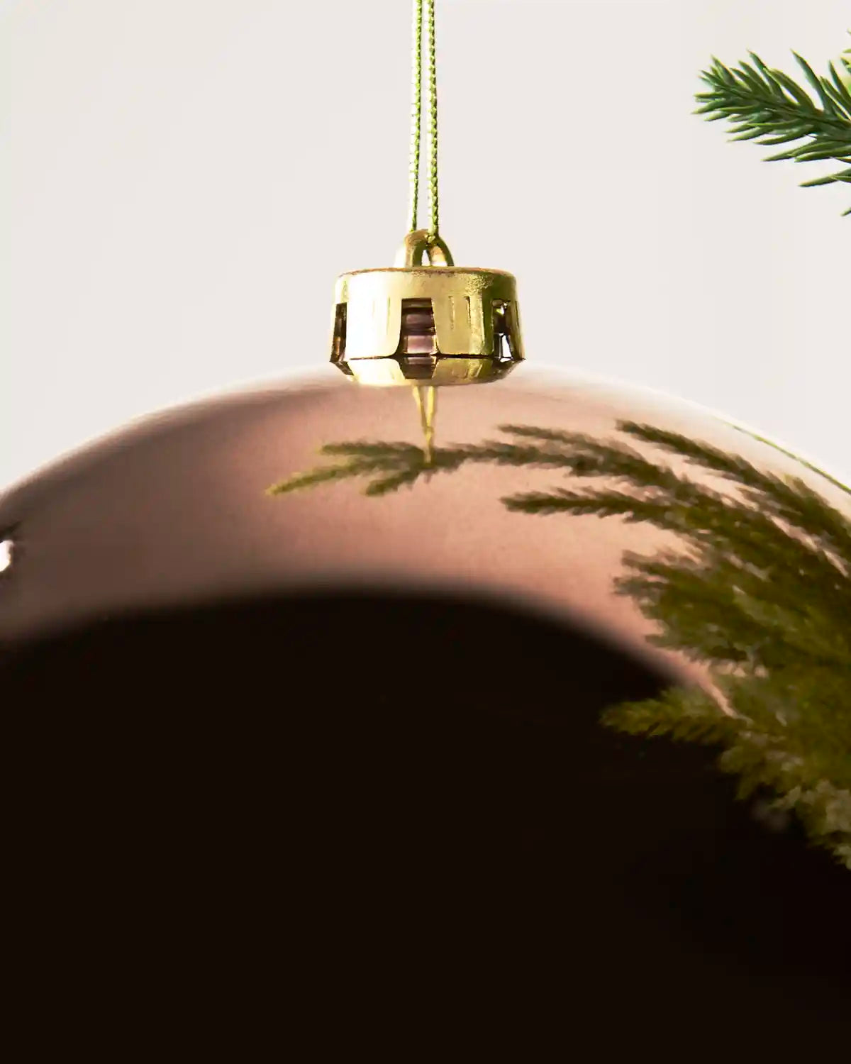 A close-up of a glossy Mocha Christmas ornament with a gold hanging cap and string, reflecting pine needles against a light background.