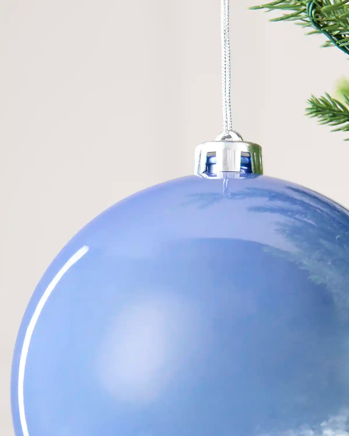 A close-up of a glossy light blue Christmas ornament hanging from a silver cap with a white string, with a small green pine branch visible at the top right.