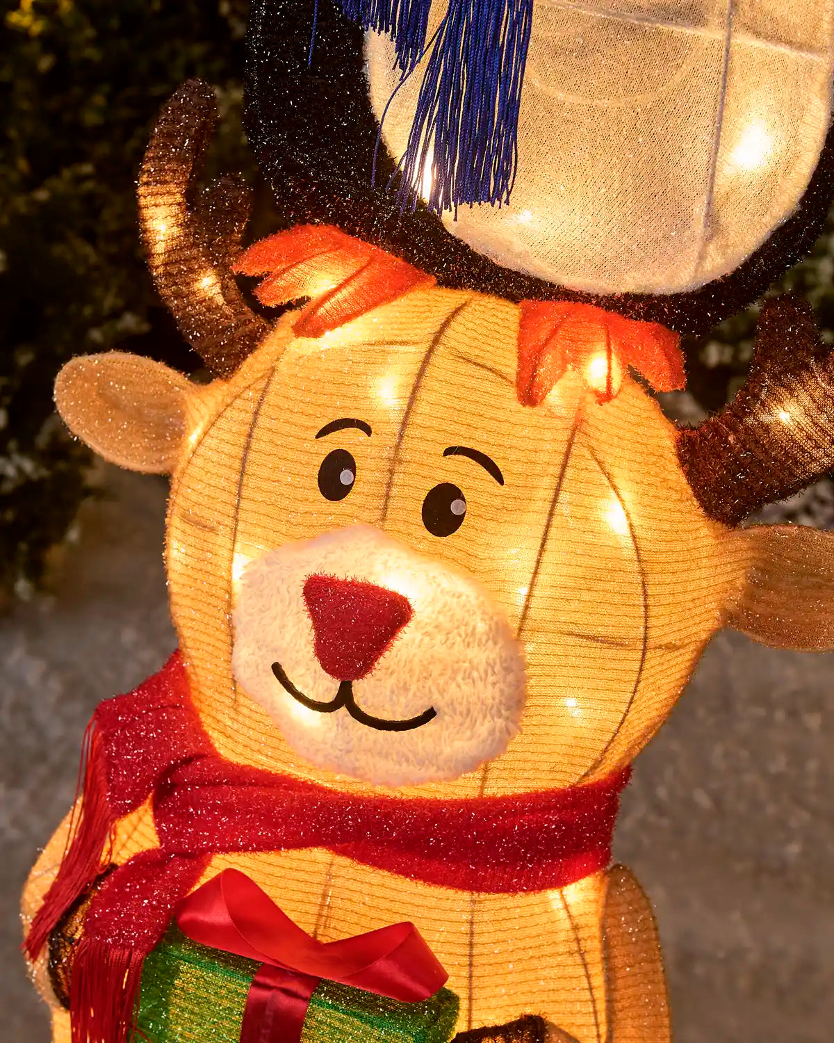 A close-up of a glowing Christmas reindeer decoration with warm white string lights, a red glittery scarf, a red nose, brown antlers, and a green gift box at the bottom.